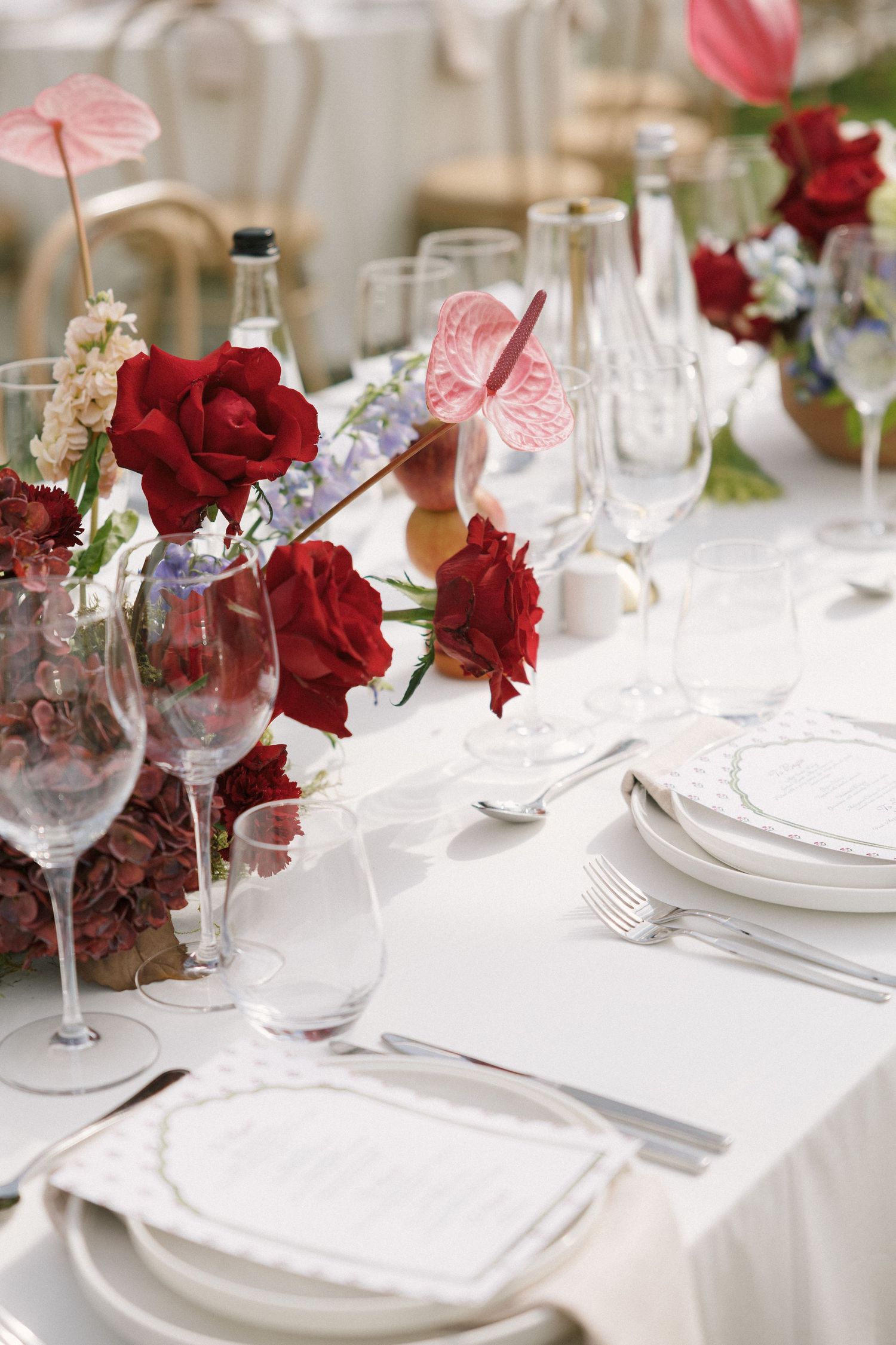 Elegant wedding reception table set with red roses, crystal wine glasses, white plates, and silverware on a white tablecloth.