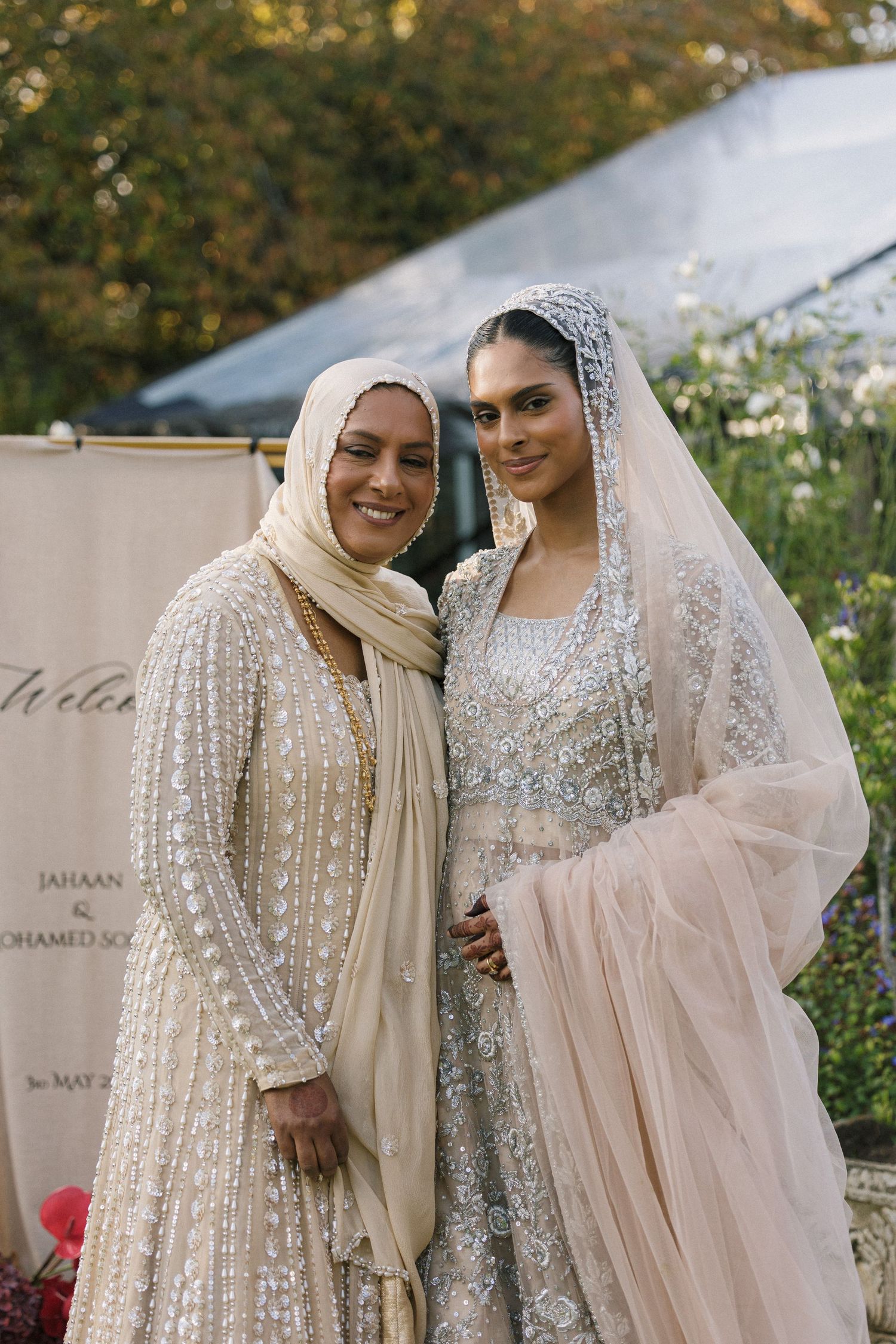 Bride in silver embroidered lehenga with veil poses with woman in beige hijab at South Asian wedding.