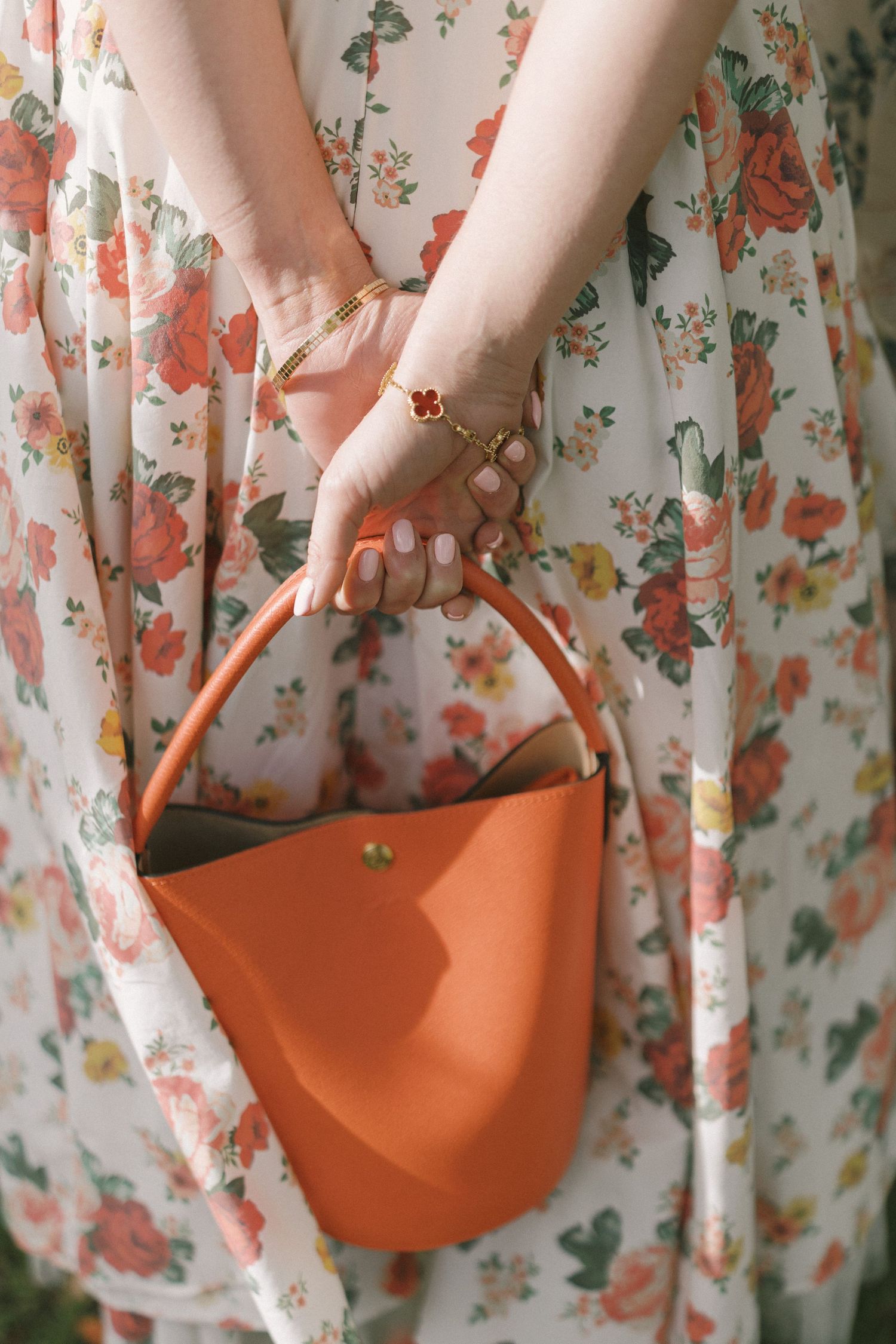 A person wearing a floral dress holds a small orange crescent-shaped handbag against a floral background.