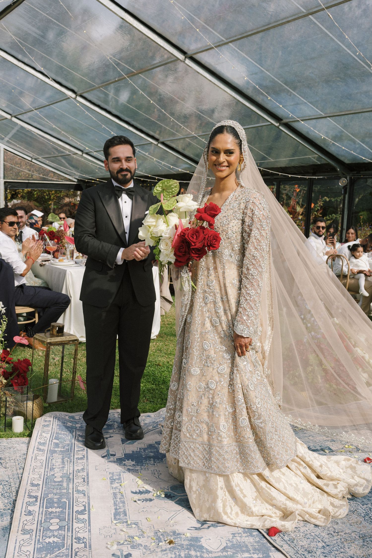 Bride in embellished ivory lehenga with veil and groom in black tuxedo stand together at outdoor wedding ceremony.