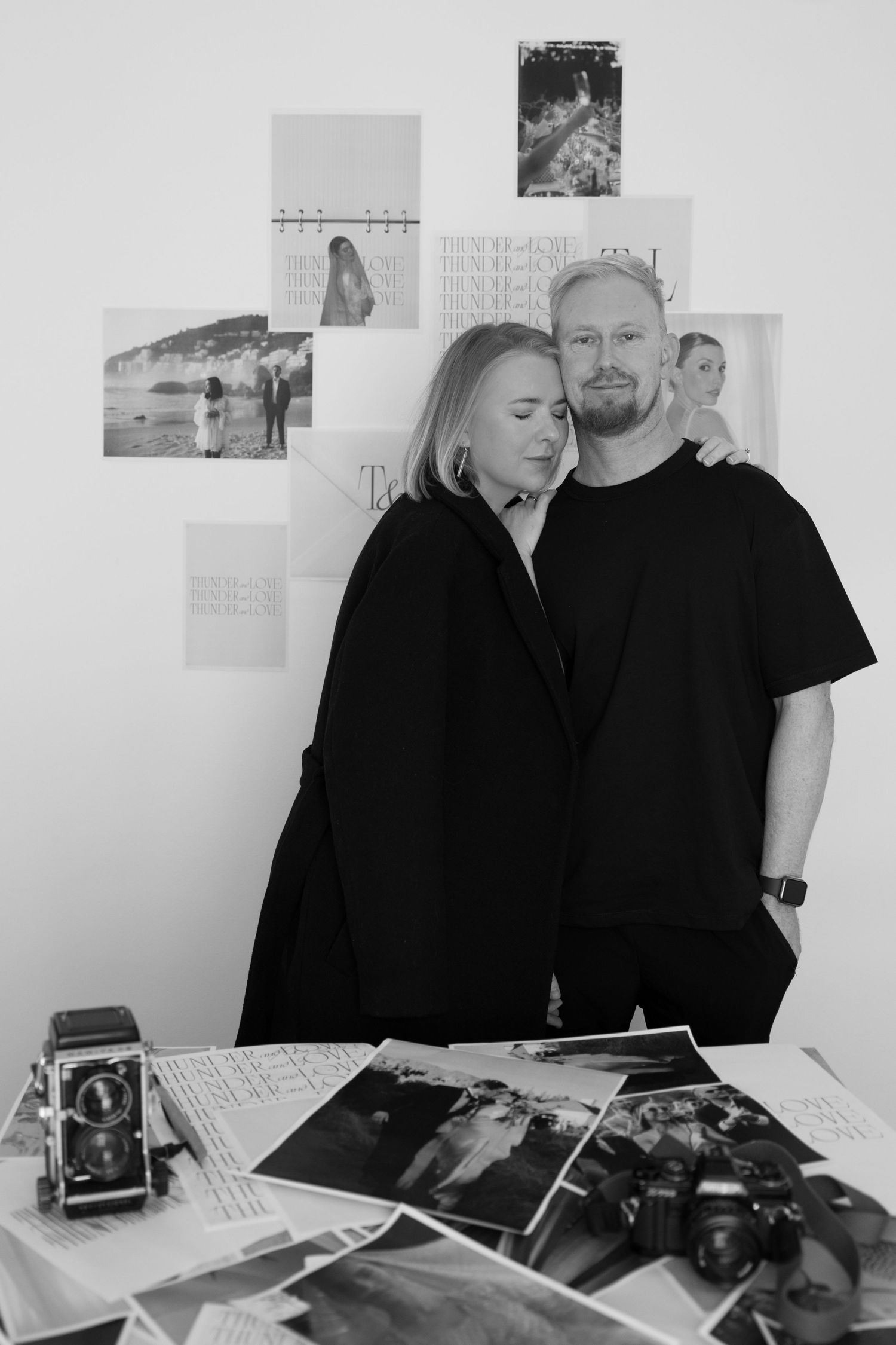 Two people in black clothing look through photographs together at a desk with photos and calendars on white wall.