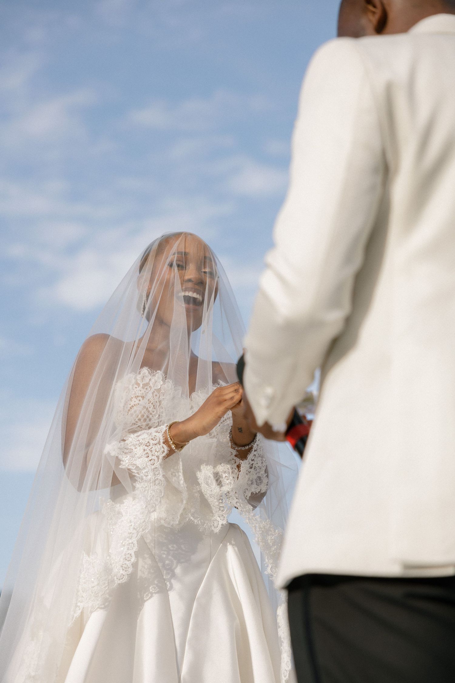 A bride in a white lace gown and veil holds hands with a groom in a white suit under a blue sky.