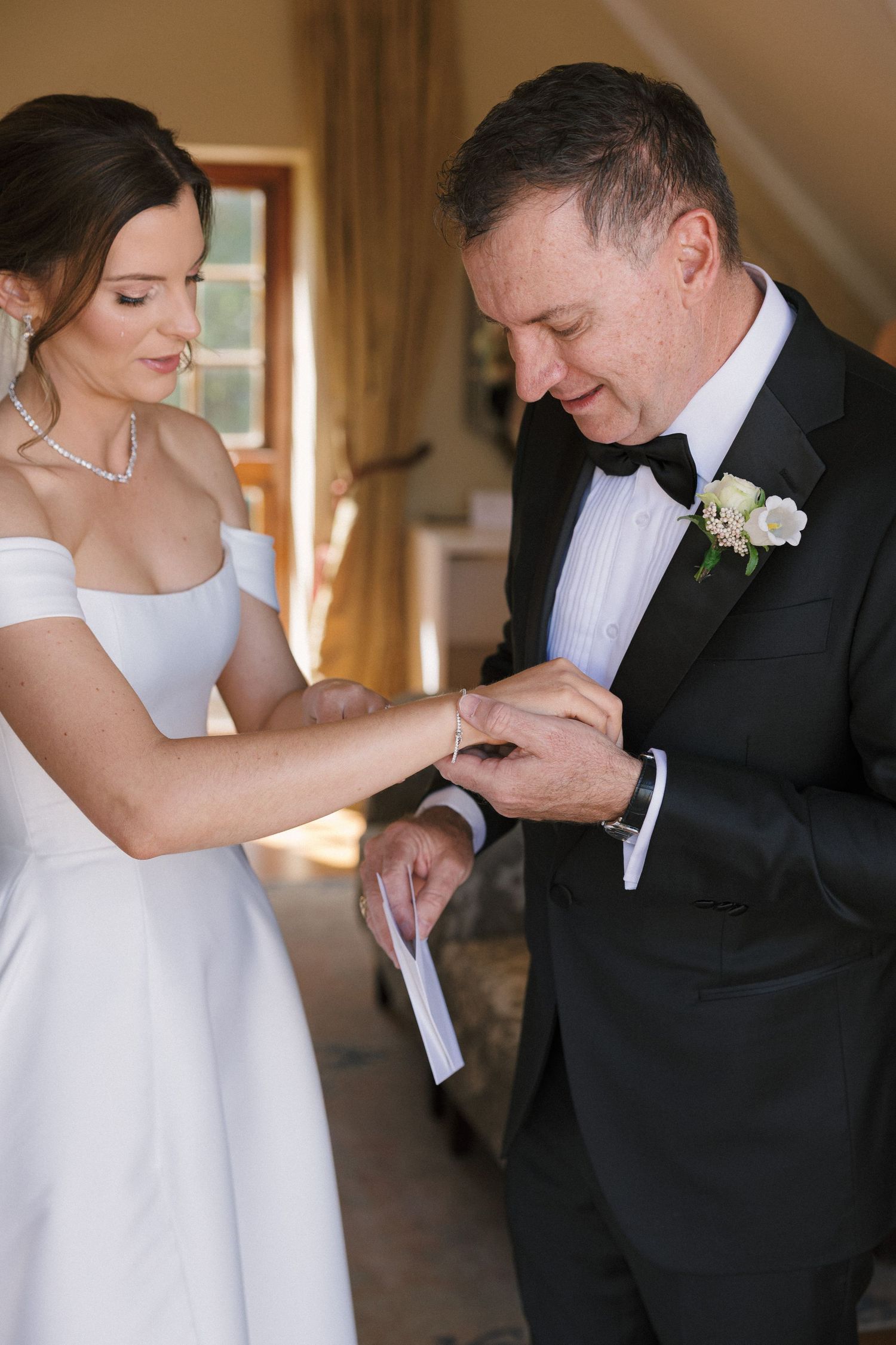 A tender moment at a wedding as the bride helps the groom with his boutonniere in a sunlit hallway.