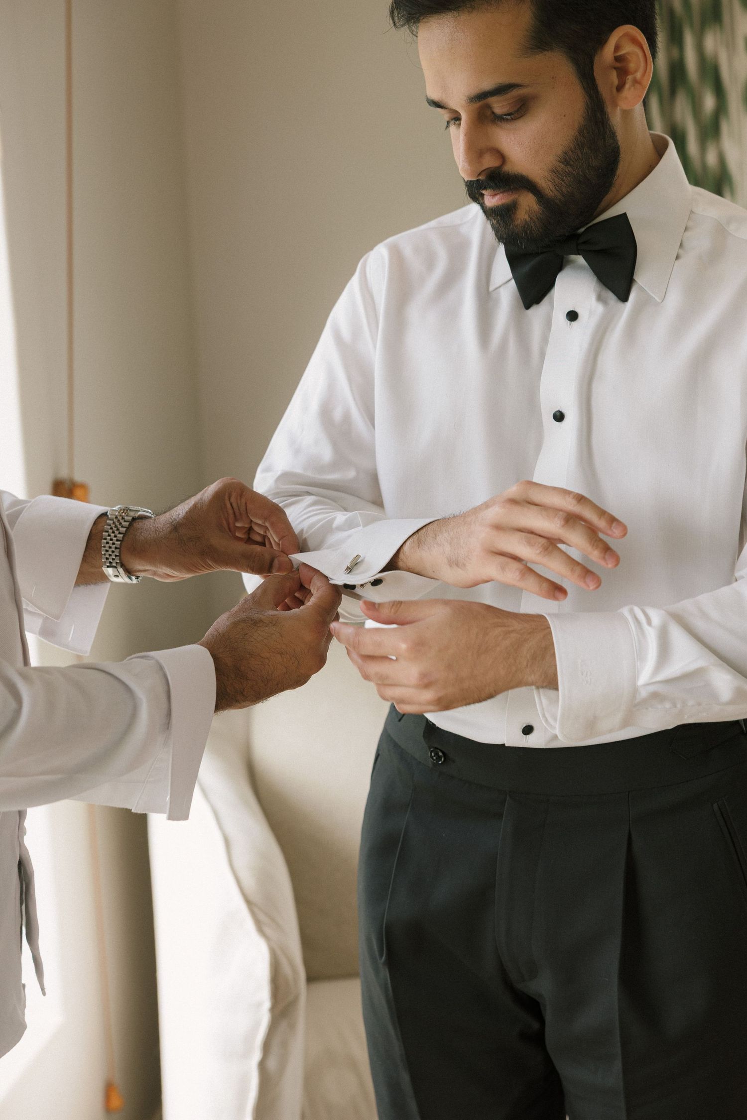 A bearded groom in a white tuxedo shirt and bow tie gets his cufflinks fastened by a helper while preparing for his wedding.