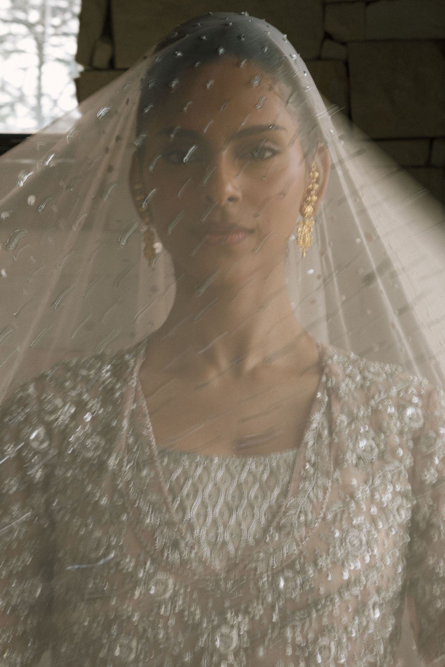Bride wearing an embellished white gown and pearl-dotted veil with gold drop earrings in a soft editorial portrait.