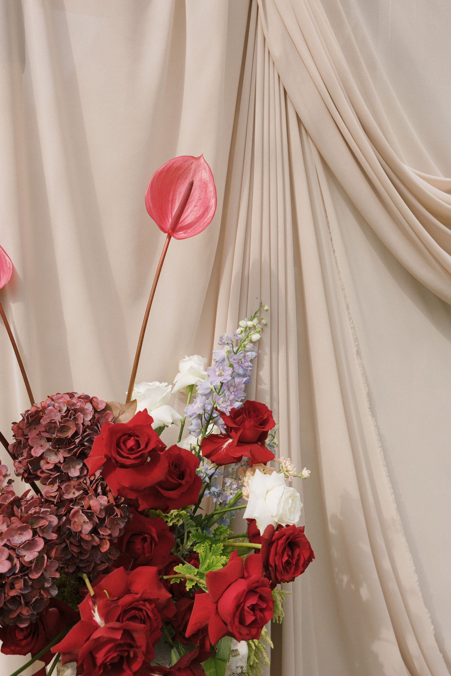 Elegant floral arrangement with red roses, pink anthuriums, and hydrangeas against a draped beige fabric backdrop.