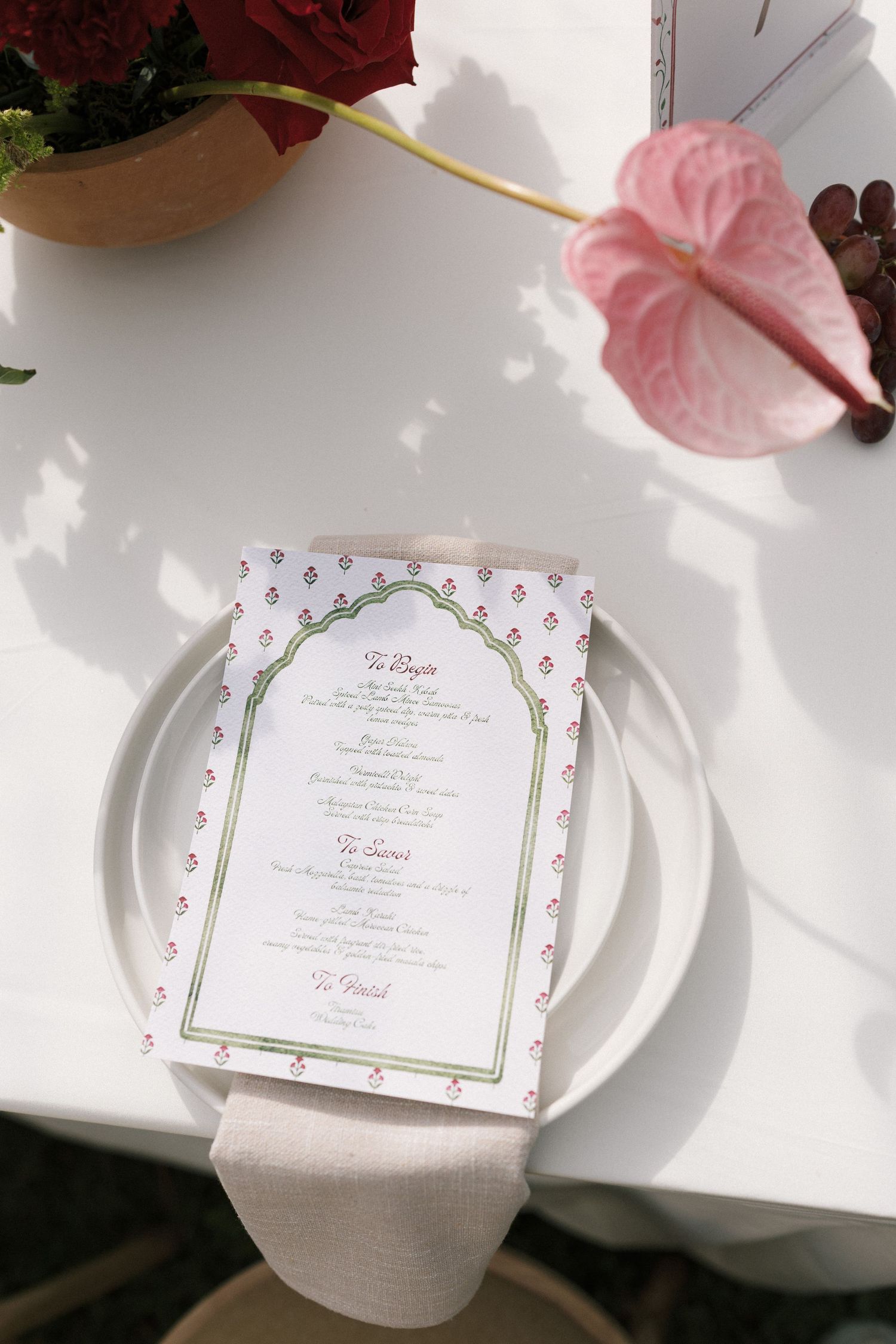 Elegant wedding place setting with floral menu card on white plate, pink anthurium flower and red roses on white tablecloth.