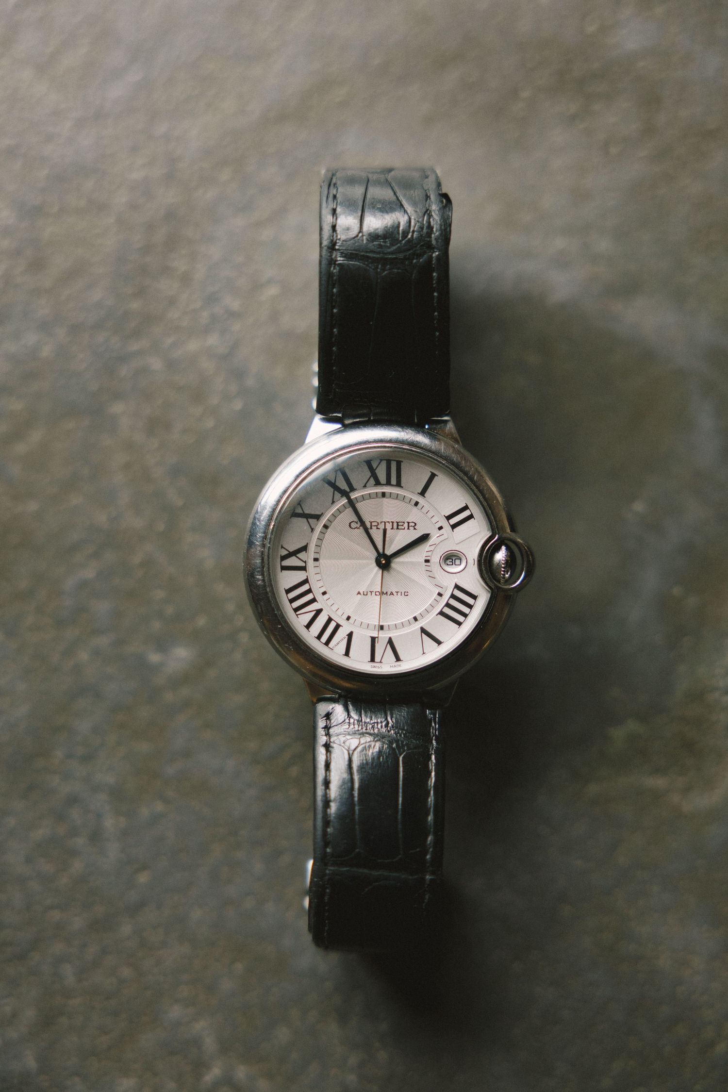 A luxury Cartier Ballon Bleu watch with silver dial and black leather strap displayed on a textured gray surface.