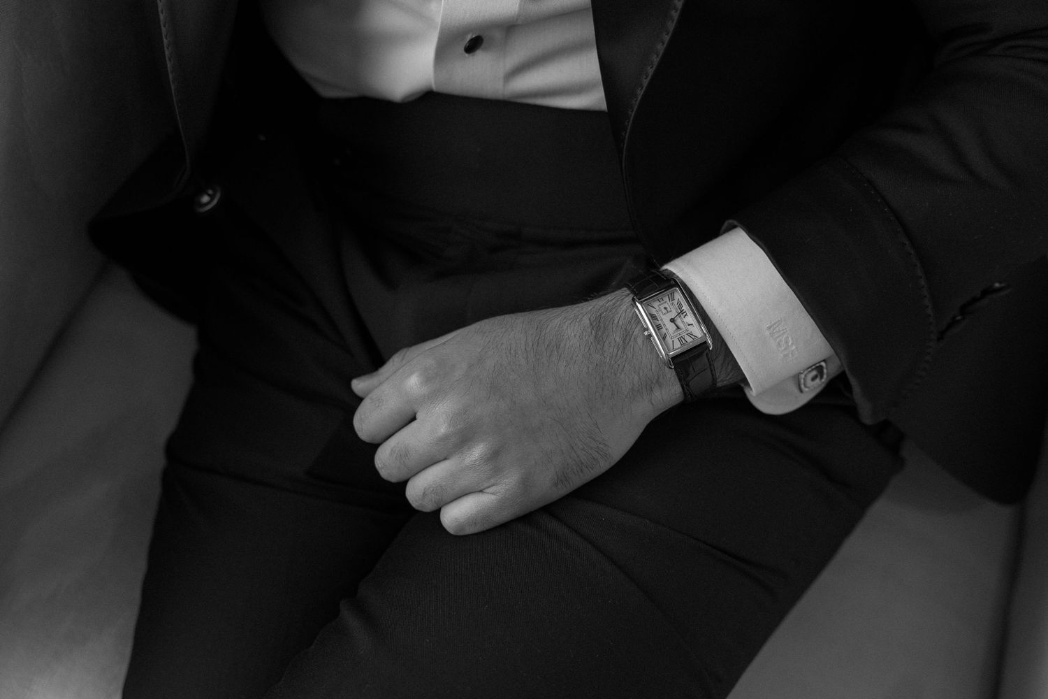 Black and white photo of a man in a tuxedo suit displaying a luxury wristwatch with elegant cufflinks.