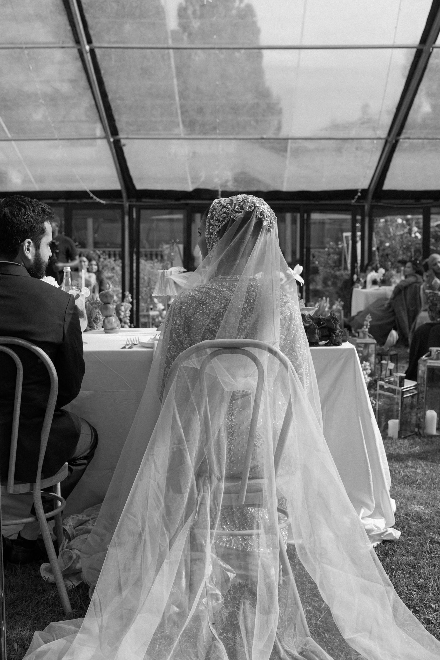 Black and white photo of a bride in a lace veil seated at a wedding ceremony table with groom beside her.
