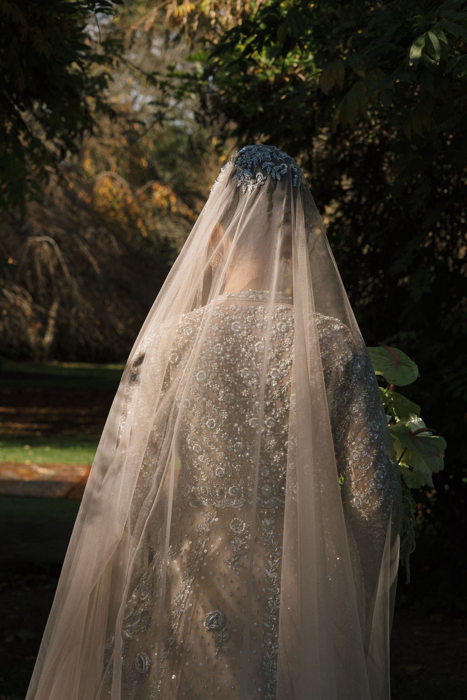 Bride seen from behind wearing an intricate beaded wedding gown and long embellished veil in a garden.