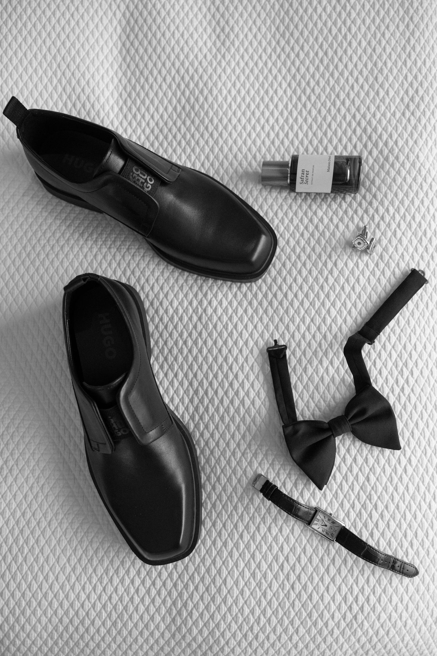 Black leather dress shoes with bow tie, cufflinks, watch, and cologne arranged on white textured surface.