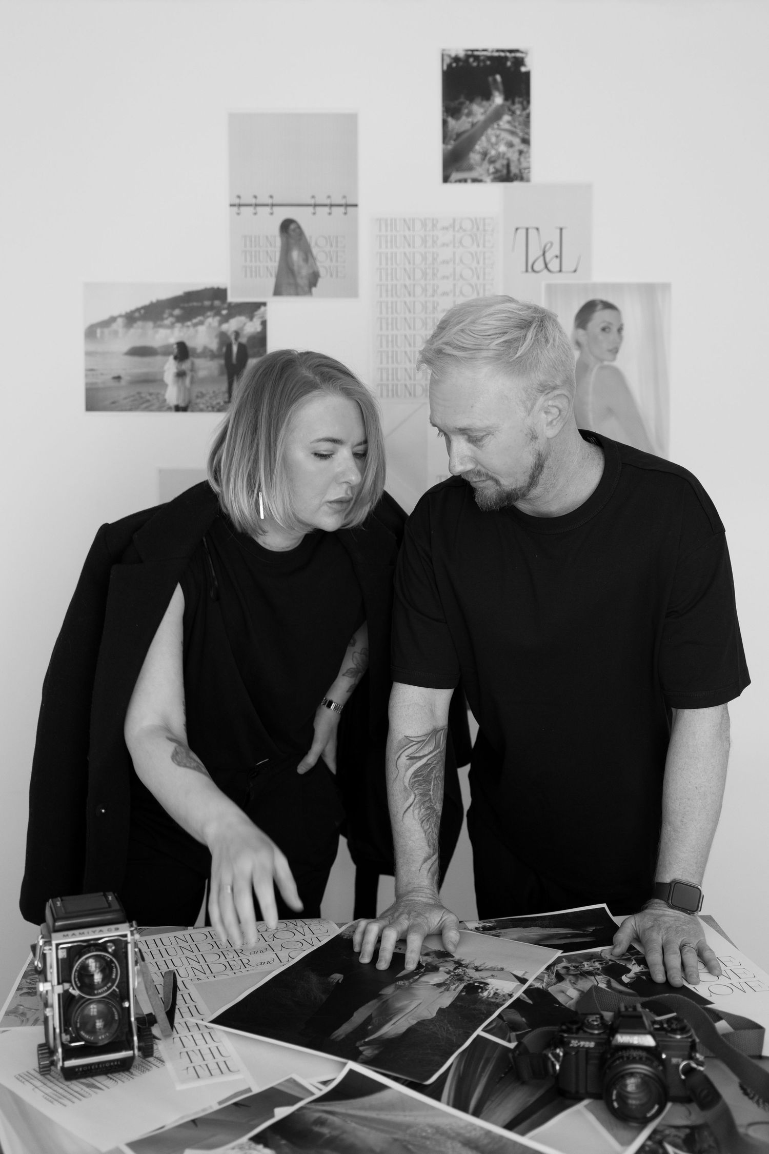 Two people in black clothing sit together at a desk looking at photographs and documents spread across the surface.