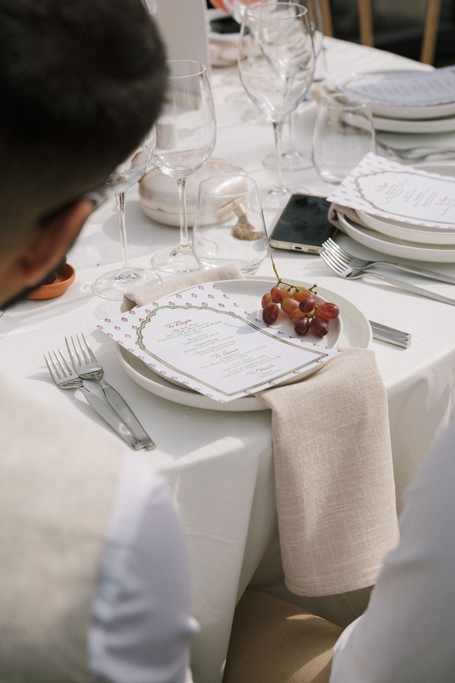 Elegant wedding reception table setting with white linens, crystal glasses, menu cards, and fresh red grapes on a plate.