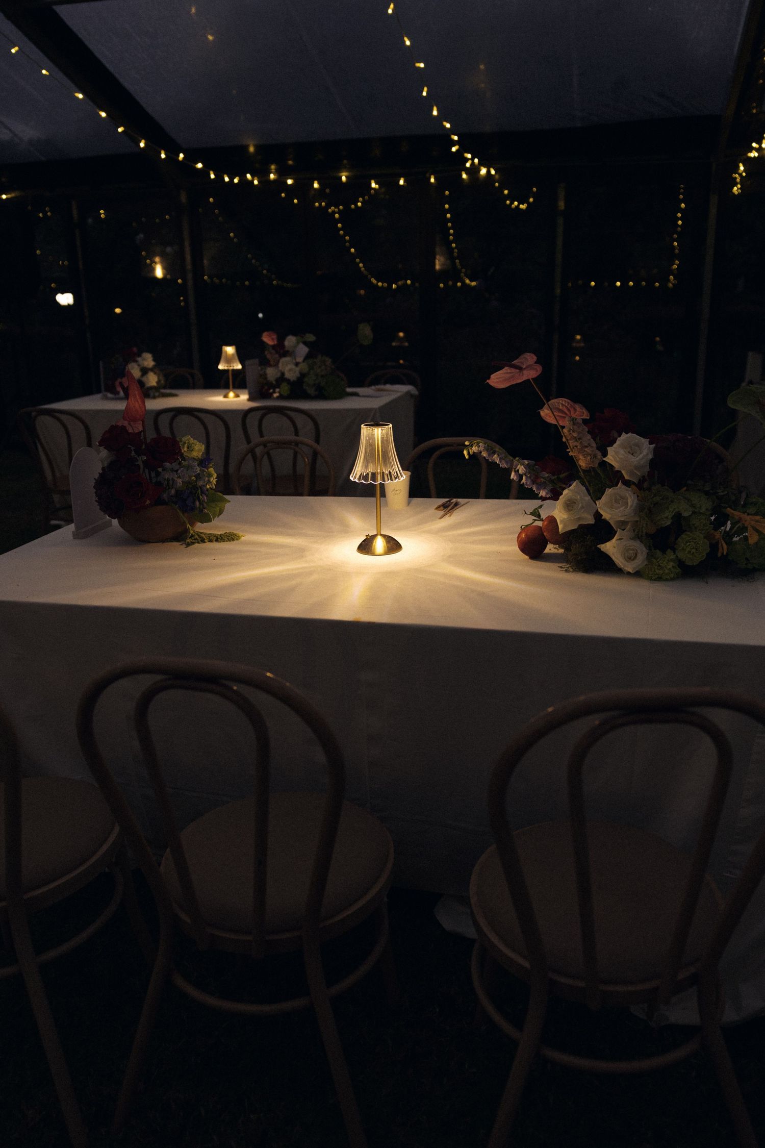 Elegant wedding reception table lit by a small lamp with floral centerpieces and string lights overhead.
