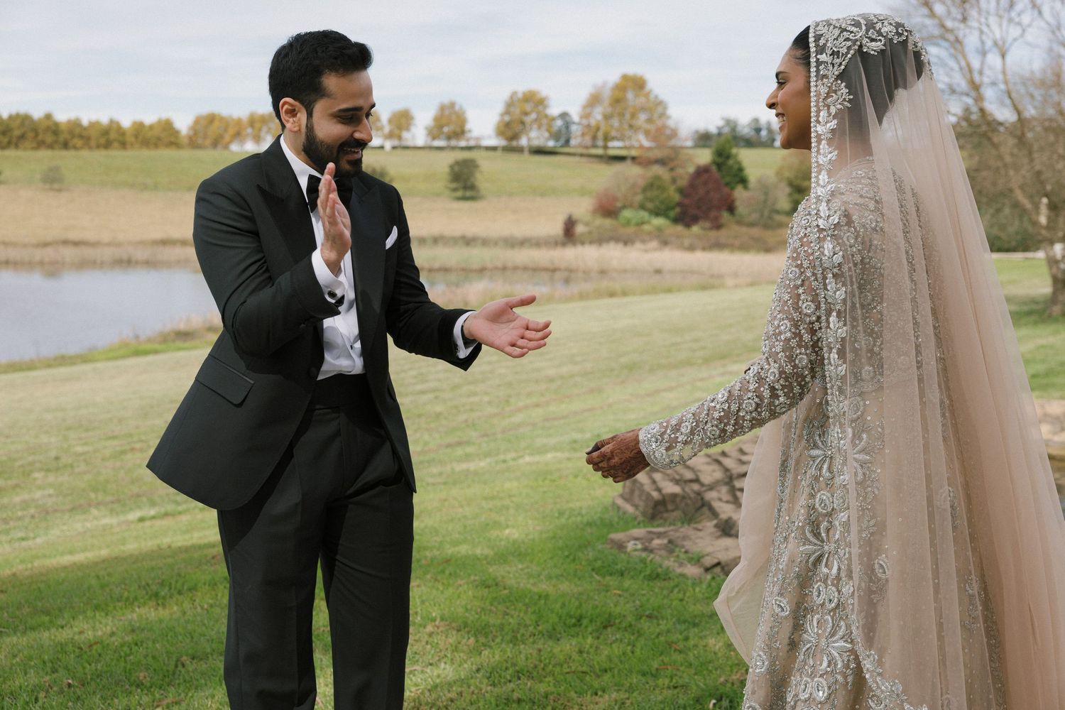 A groom in a black suit reaches out to his bride in an embellished white gown and veil at an outdoor wedding.