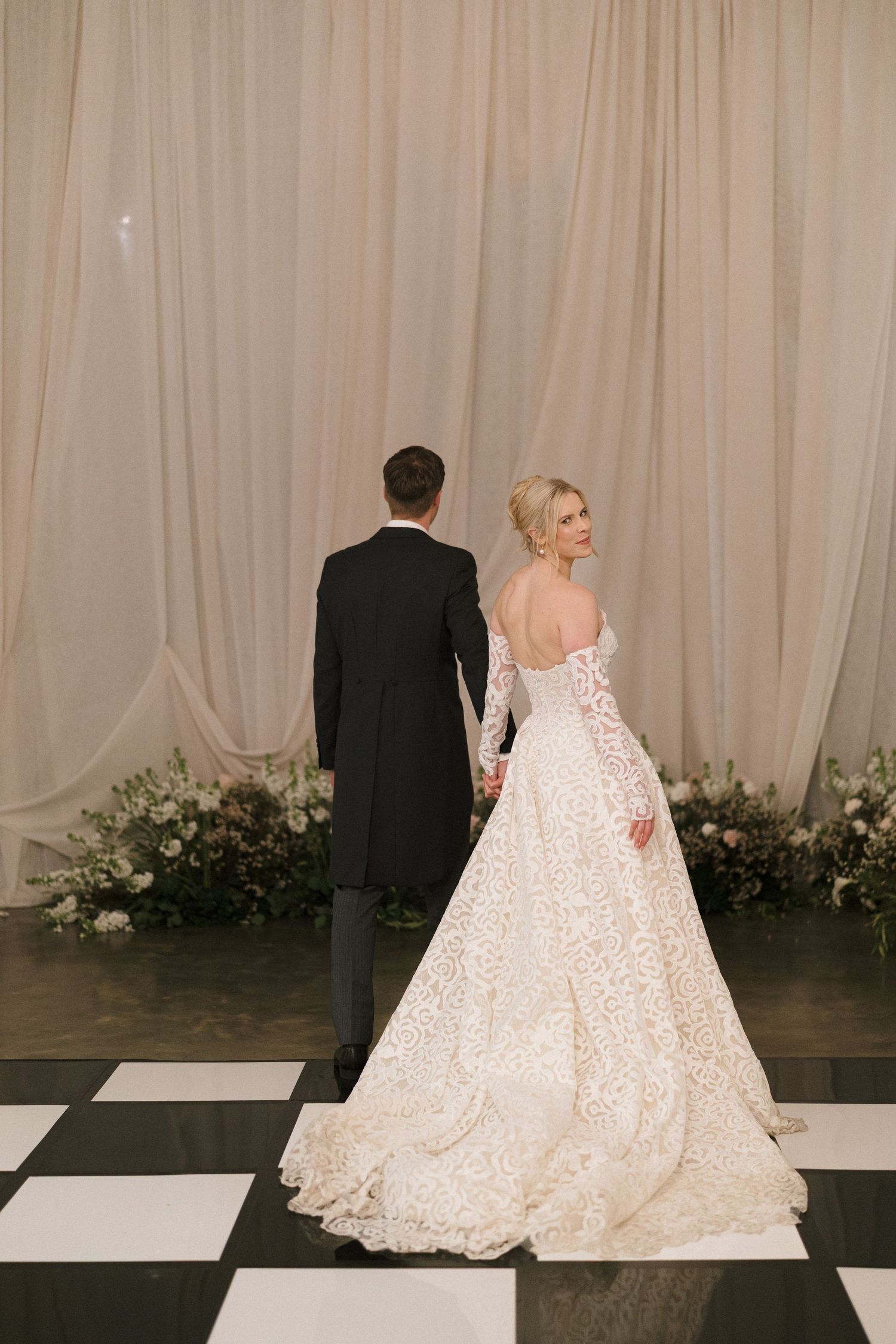 Bride in lace wedding gown and groom in black tuxedo stand on checkered floor against draped fabric and floral backdrop.