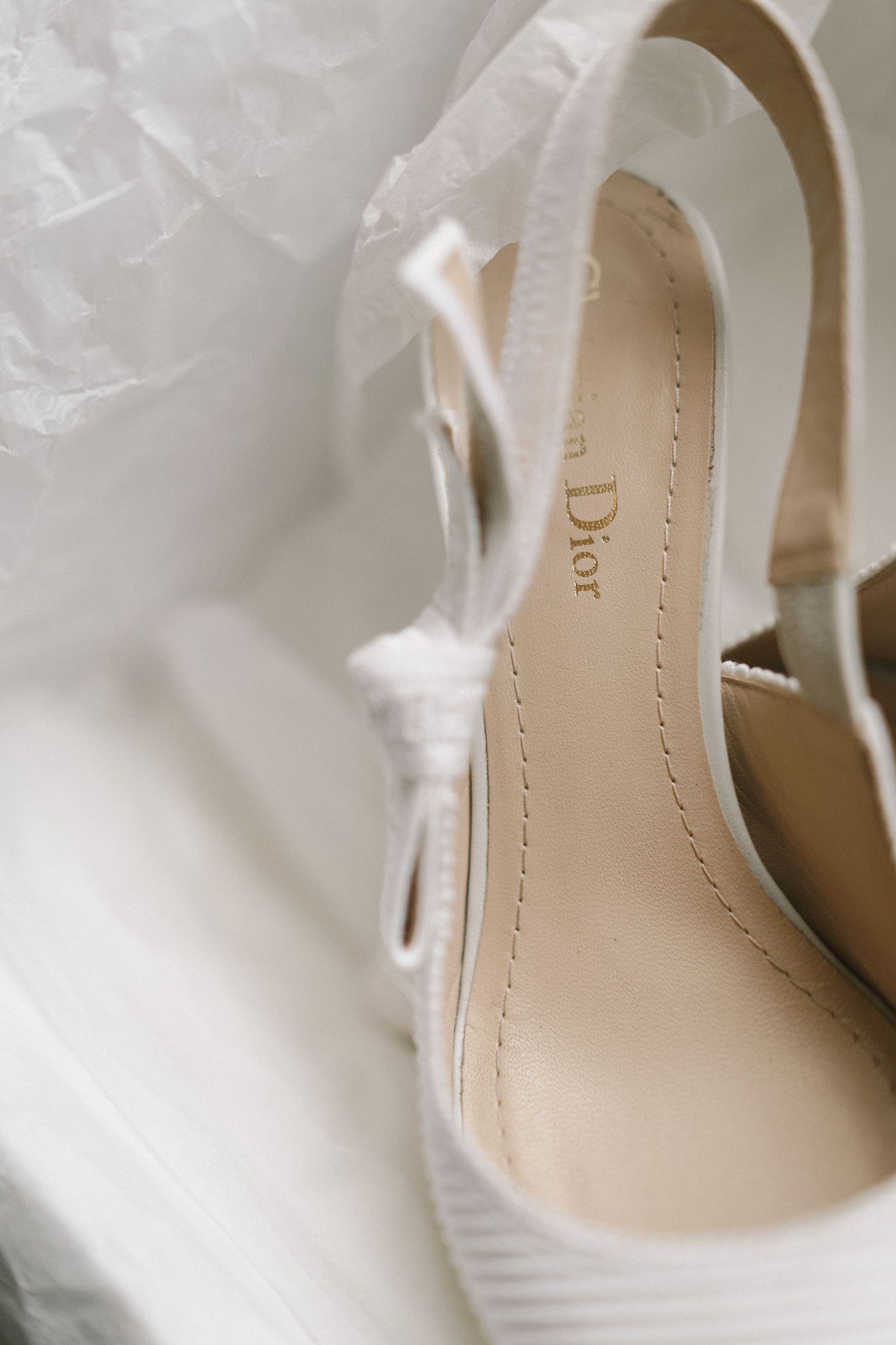 Close-up view of an elegant white satin bridal shoe with delicate detailing and curved lines against a light background.