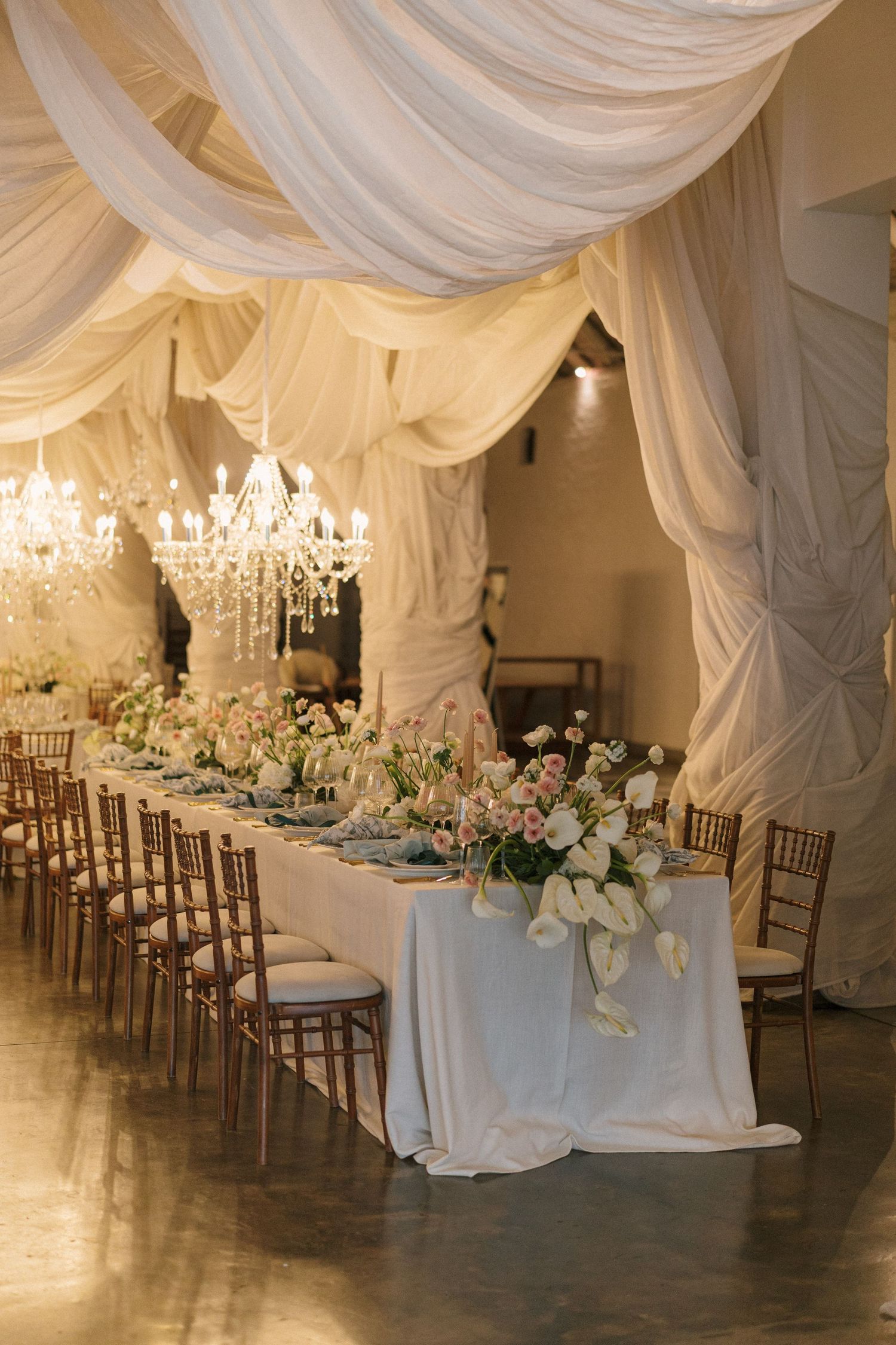 Elegant wedding reception setup with draped white fabric ceiling, crystal chandeliers, and long tables decorated with floral arrangements.