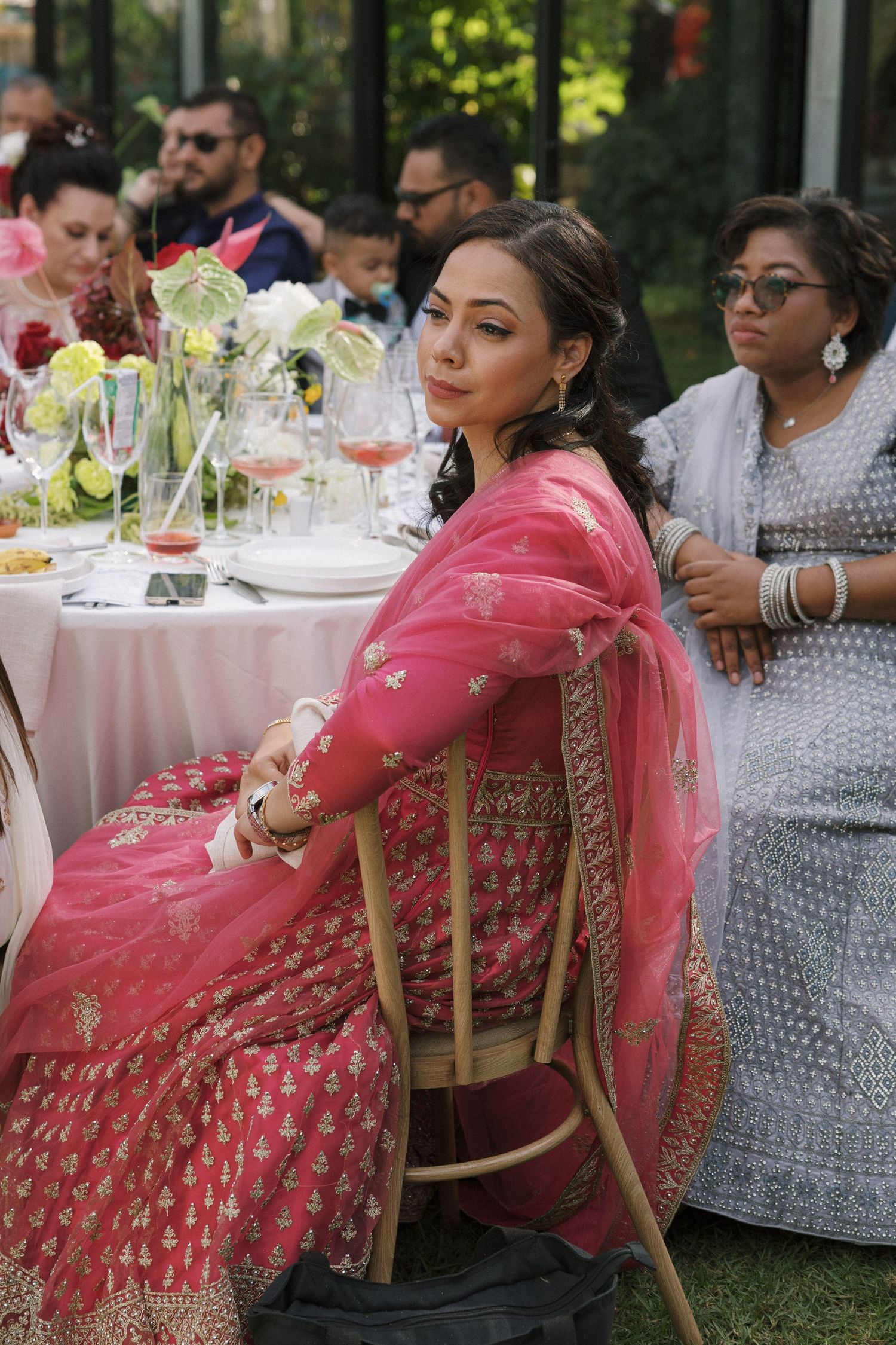 Woman in pink embroidered saree seated at outdoor wedding reception table with guests in background.