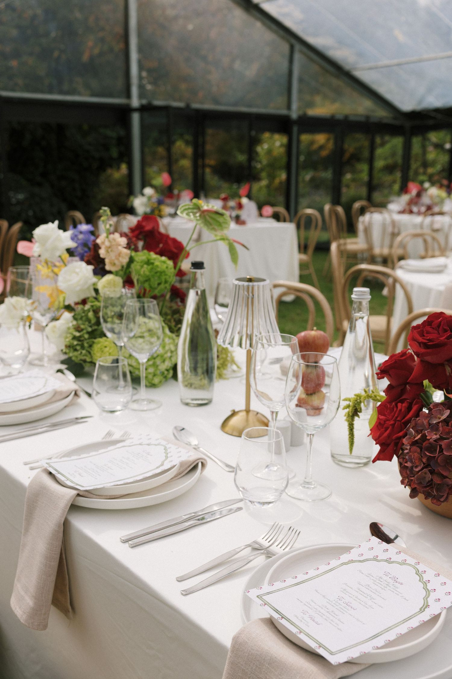 Elegant wedding reception table set with white linens, floral centerpieces, glassware, and menus in a greenhouse venue.