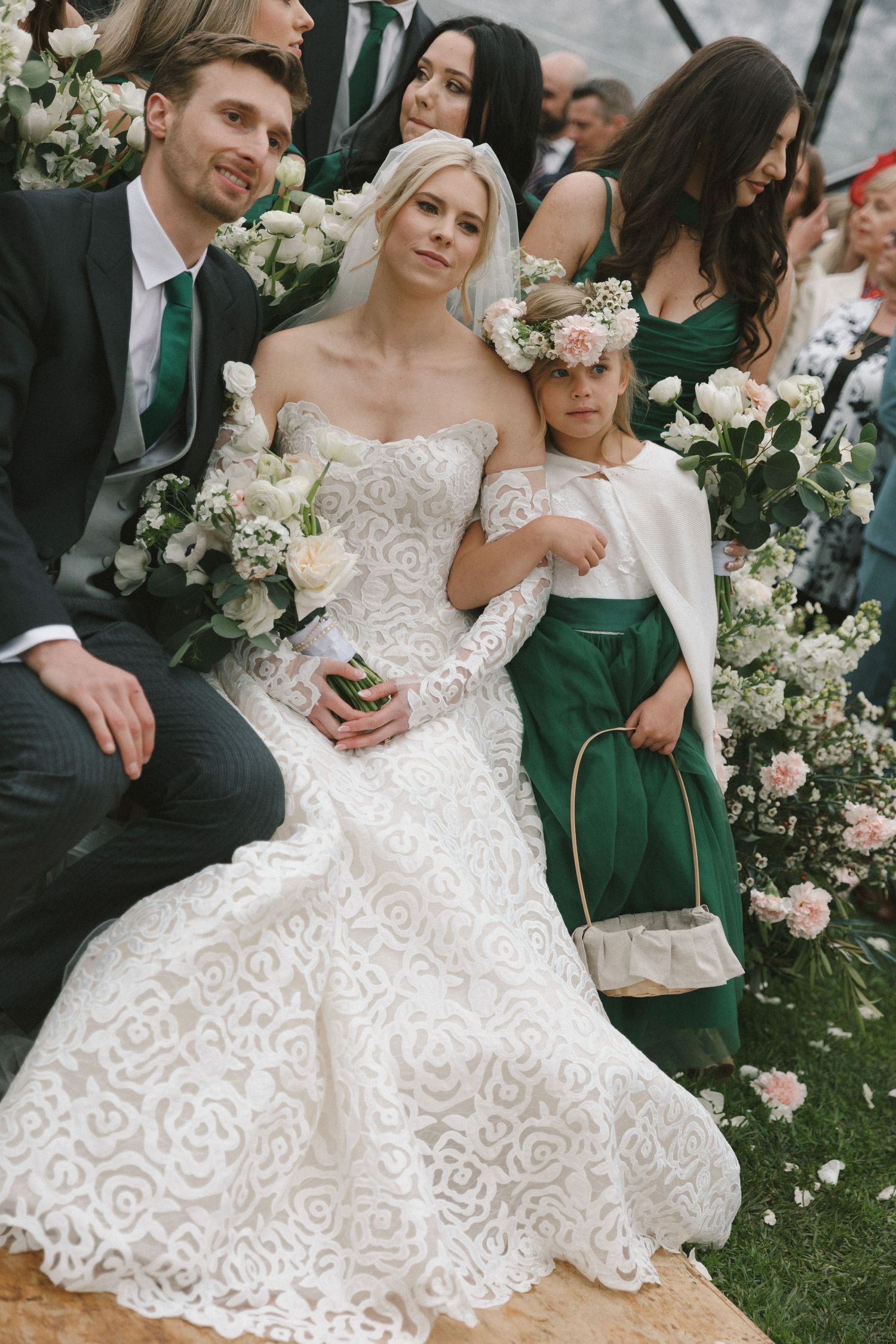 Wedding party members in formal attire gather on grass with elegant white floral arrangements and decorative elements.