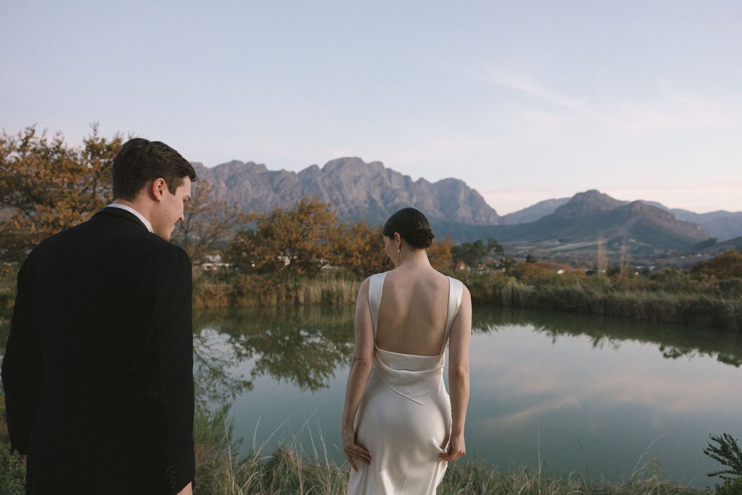 Bride and groom stand before a serene mountain lake, bride in elegant backless white gown, groom in black suit.