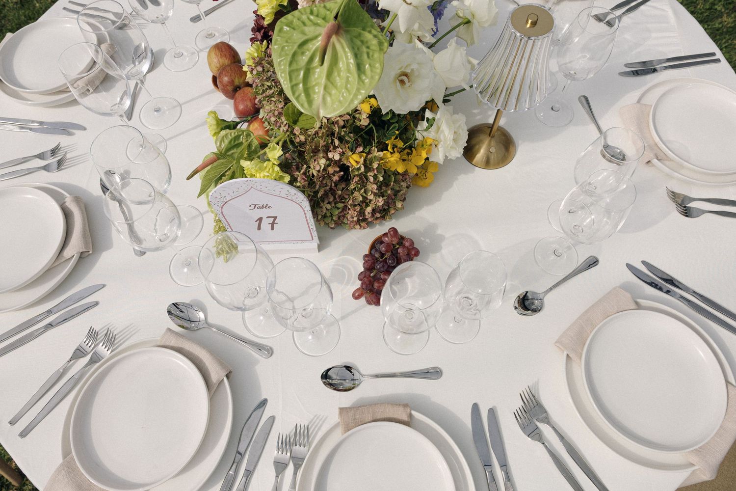 Elegant round wedding reception table with white plates, silverware, crystal glasses, and a floral centerpiece with grapes.