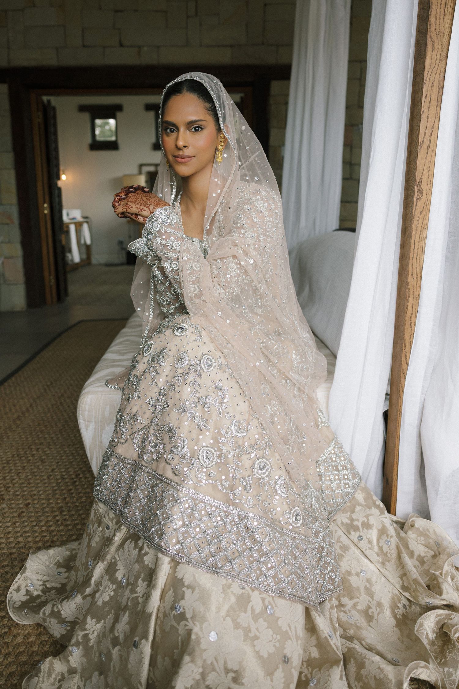 Bride in embroidered ivory lehenga with silver detailing and sheer dupatta, posing elegantly in a sunlit room.