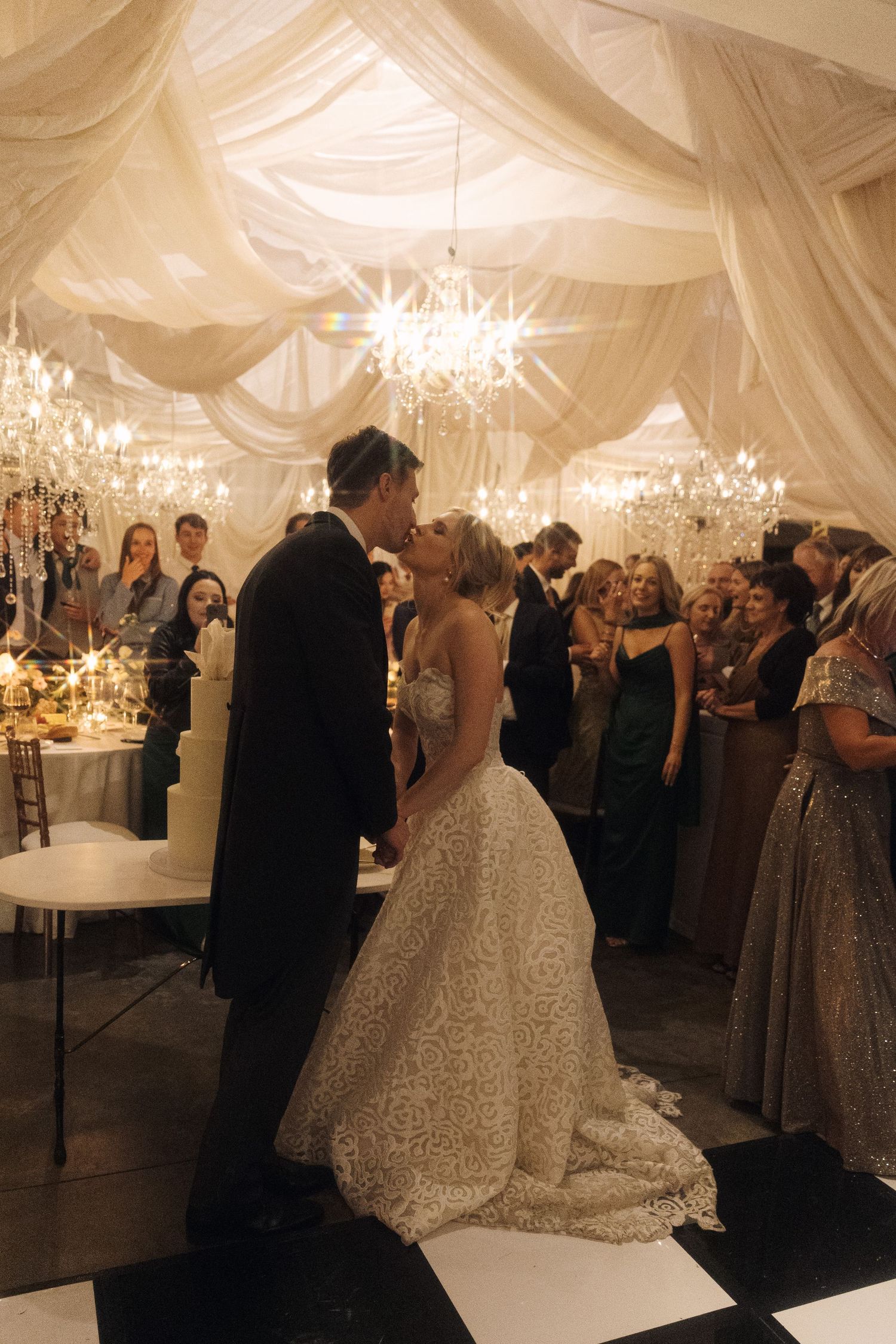Elegant wedding reception in a white draped tent with crystal chandeliers and couples dancing on the dance floor.