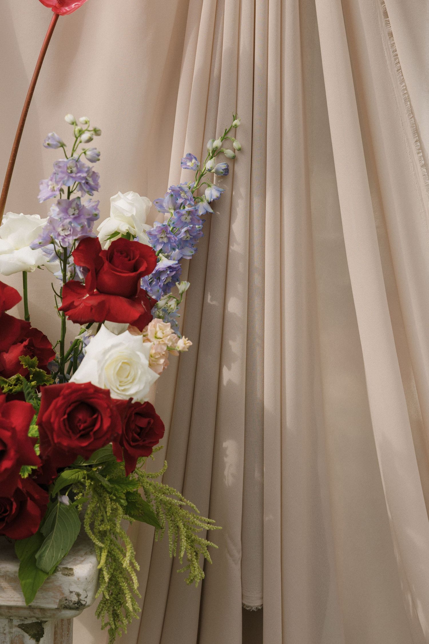 Elegant wedding floral arrangement featuring red roses, white roses, and purple delphinium against draped beige fabric backdrop.