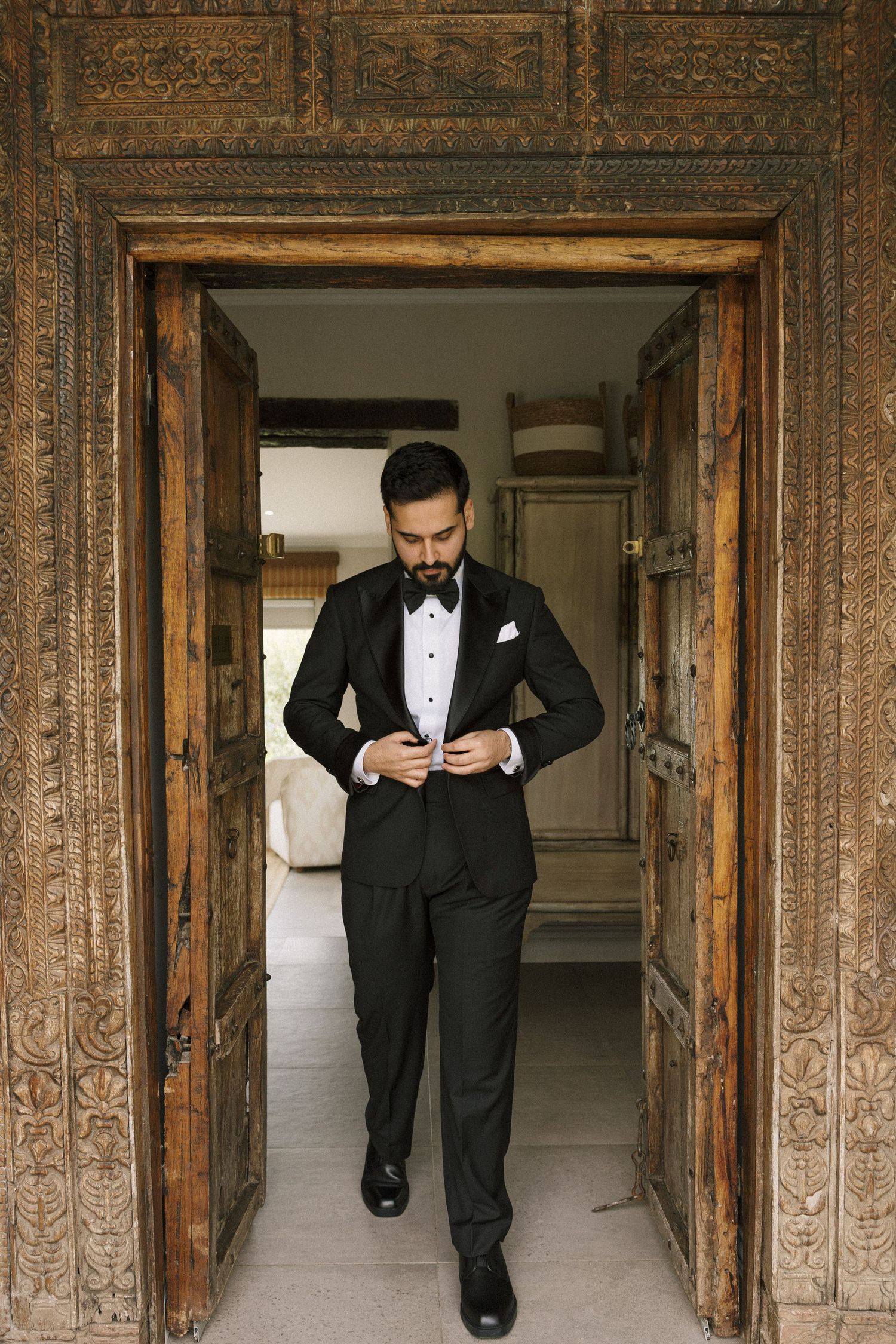 Elegant groom in black tuxedo buttoning jacket standing in ornate carved wooden doorway.