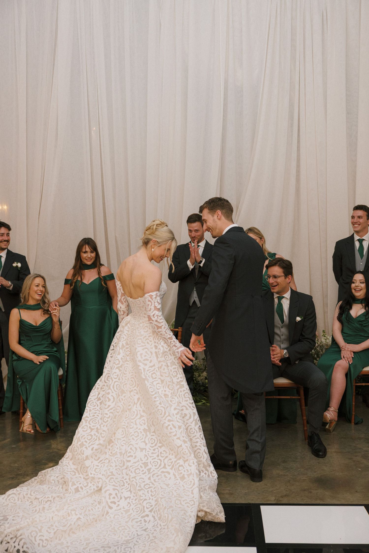 A wedding party in emerald green attire watches a couple during their first dance in front of a light backdrop.