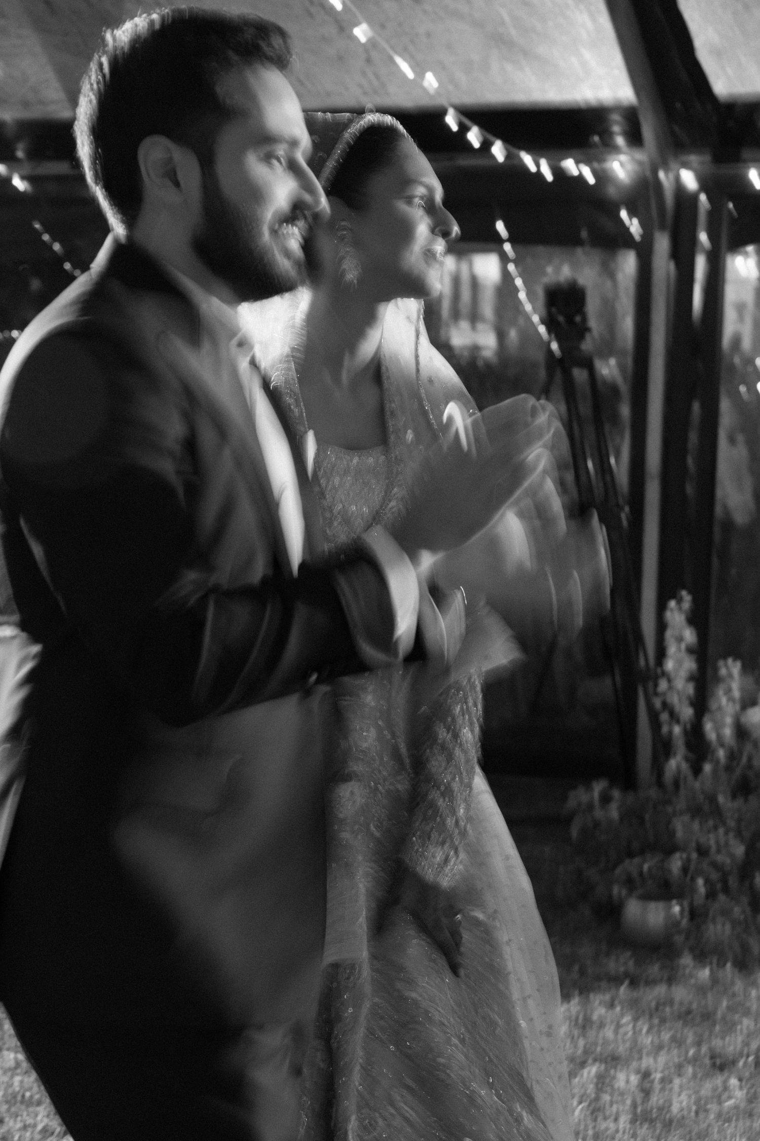 Black and white photo of a bride and groom clapping and smiling at their wedding reception under string lights.