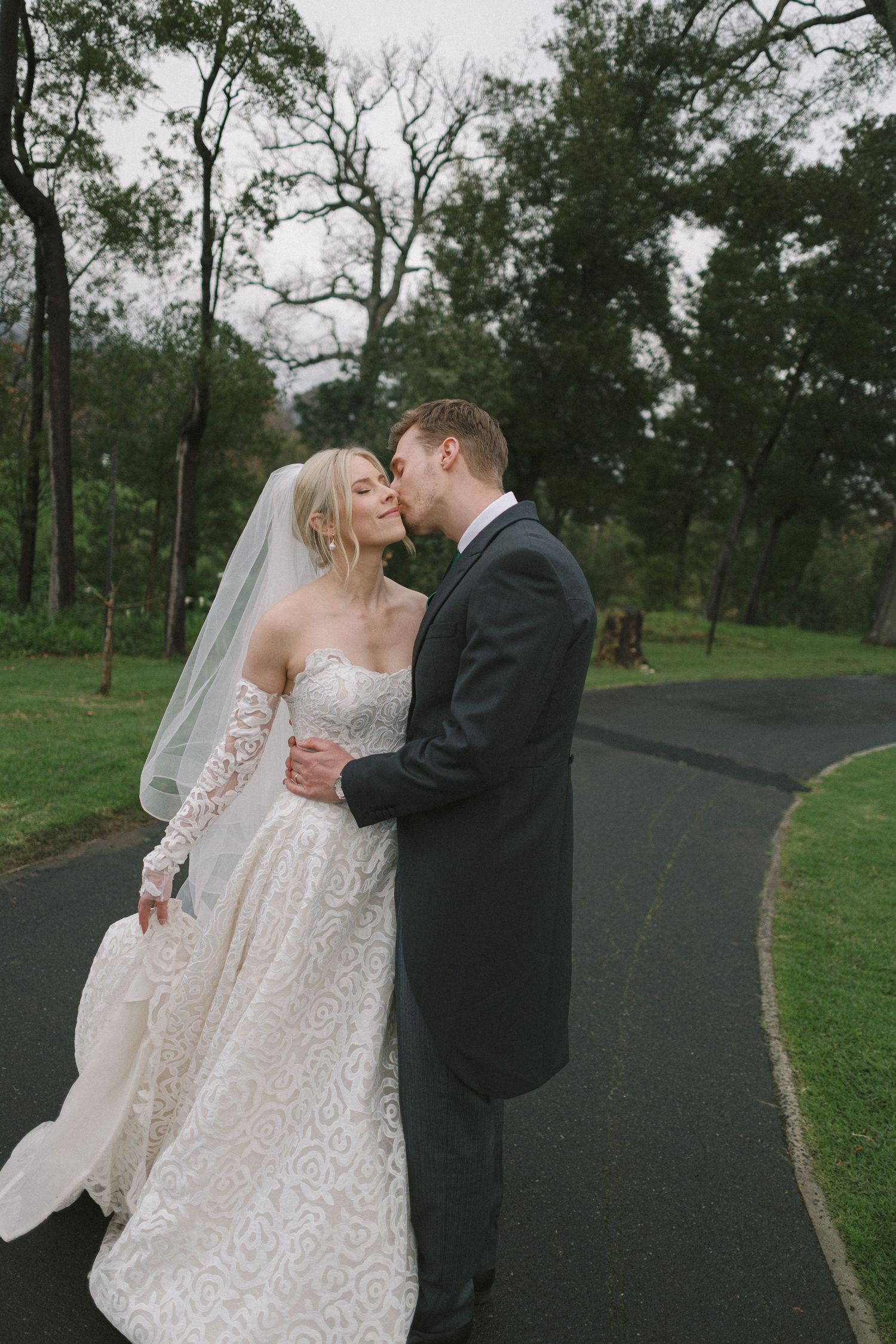 A romantic wedding couple embraces on a tree-lined path in a park setting.
