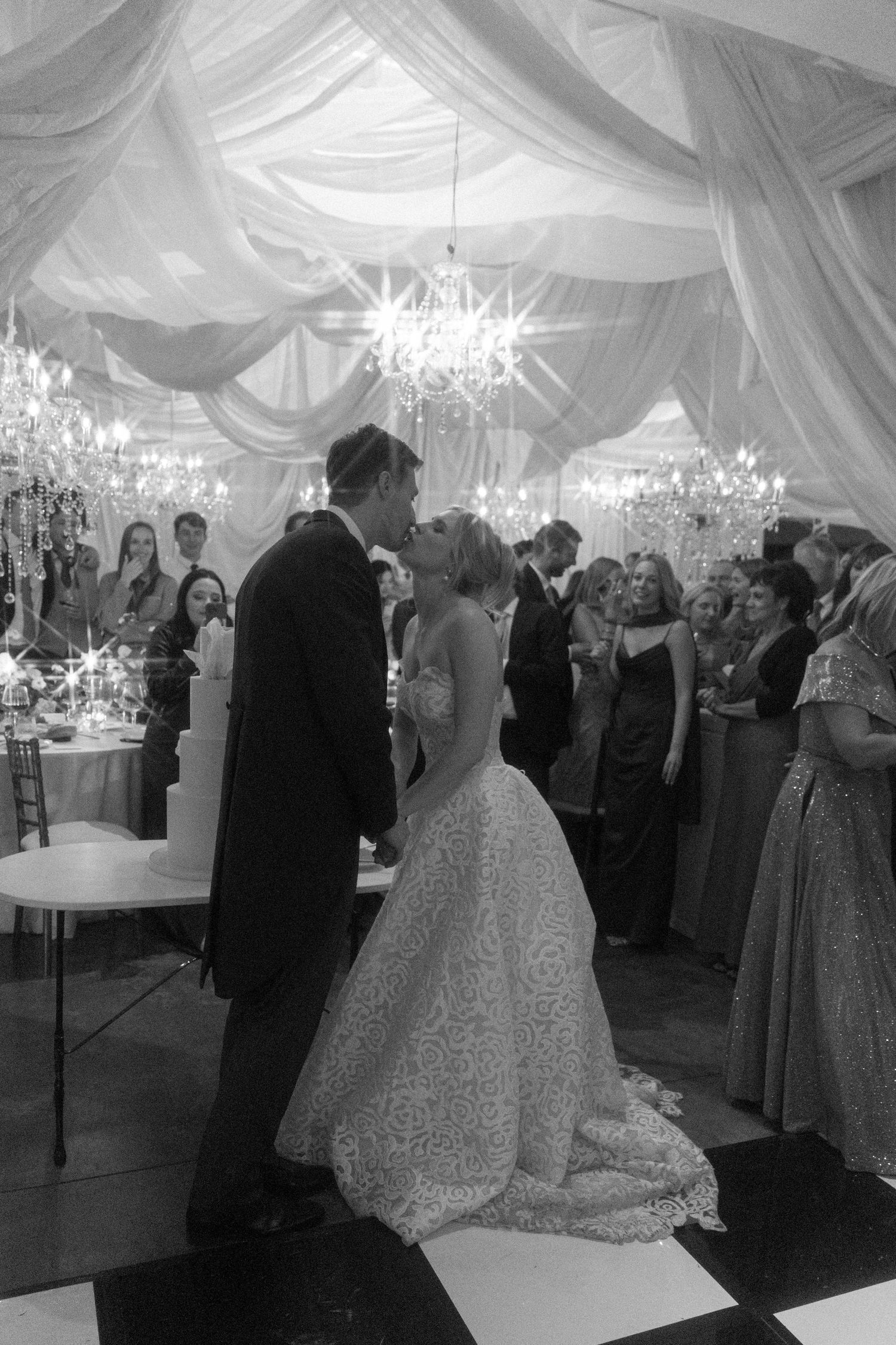 An elegant wedding reception in a white draped tent with chandeliers and guests dancing on the dance floor.