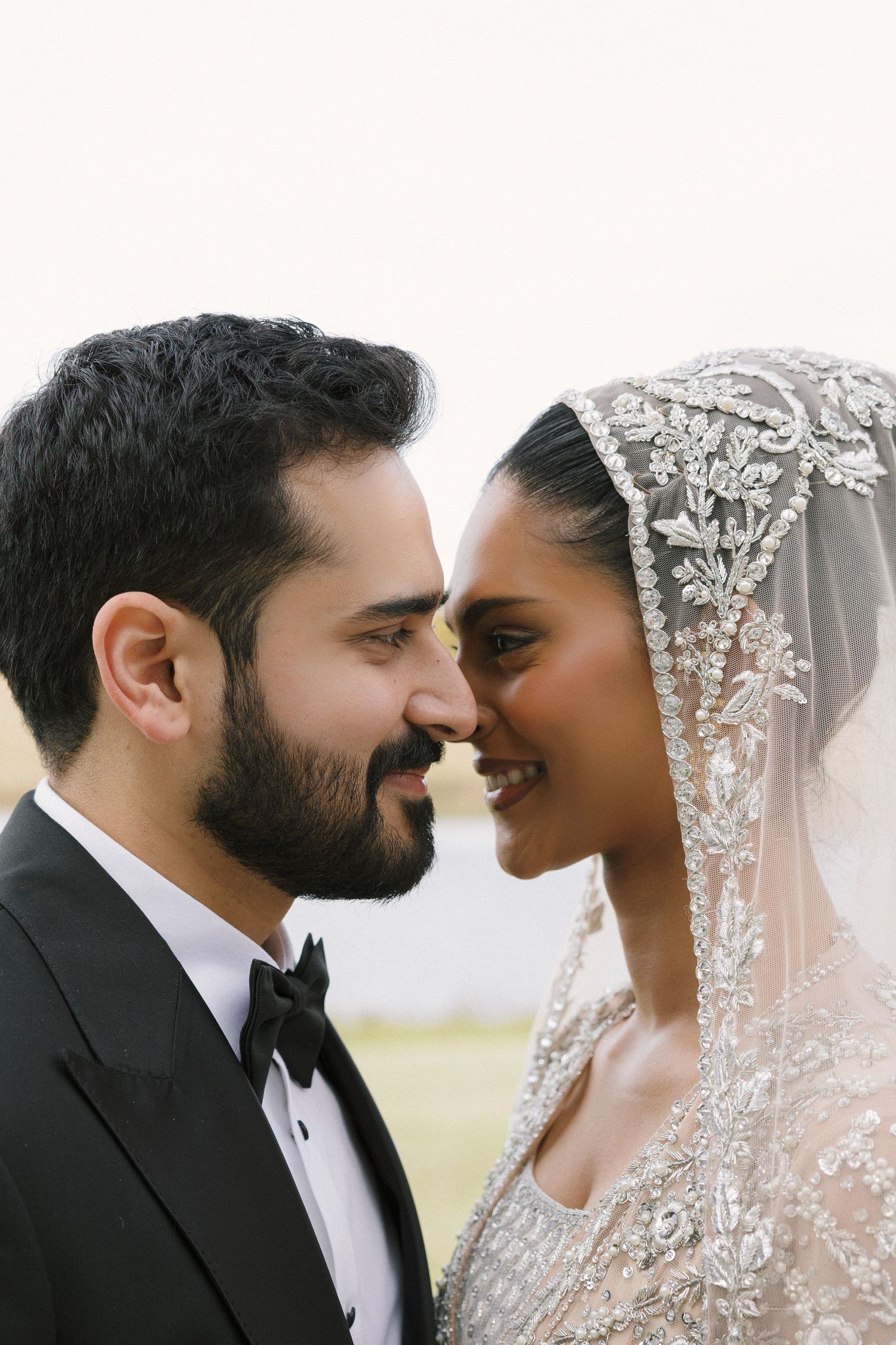 A bride in beaded veil and groom in black tuxedo share an intimate moment, foreheads nearly touching, smiling softly.