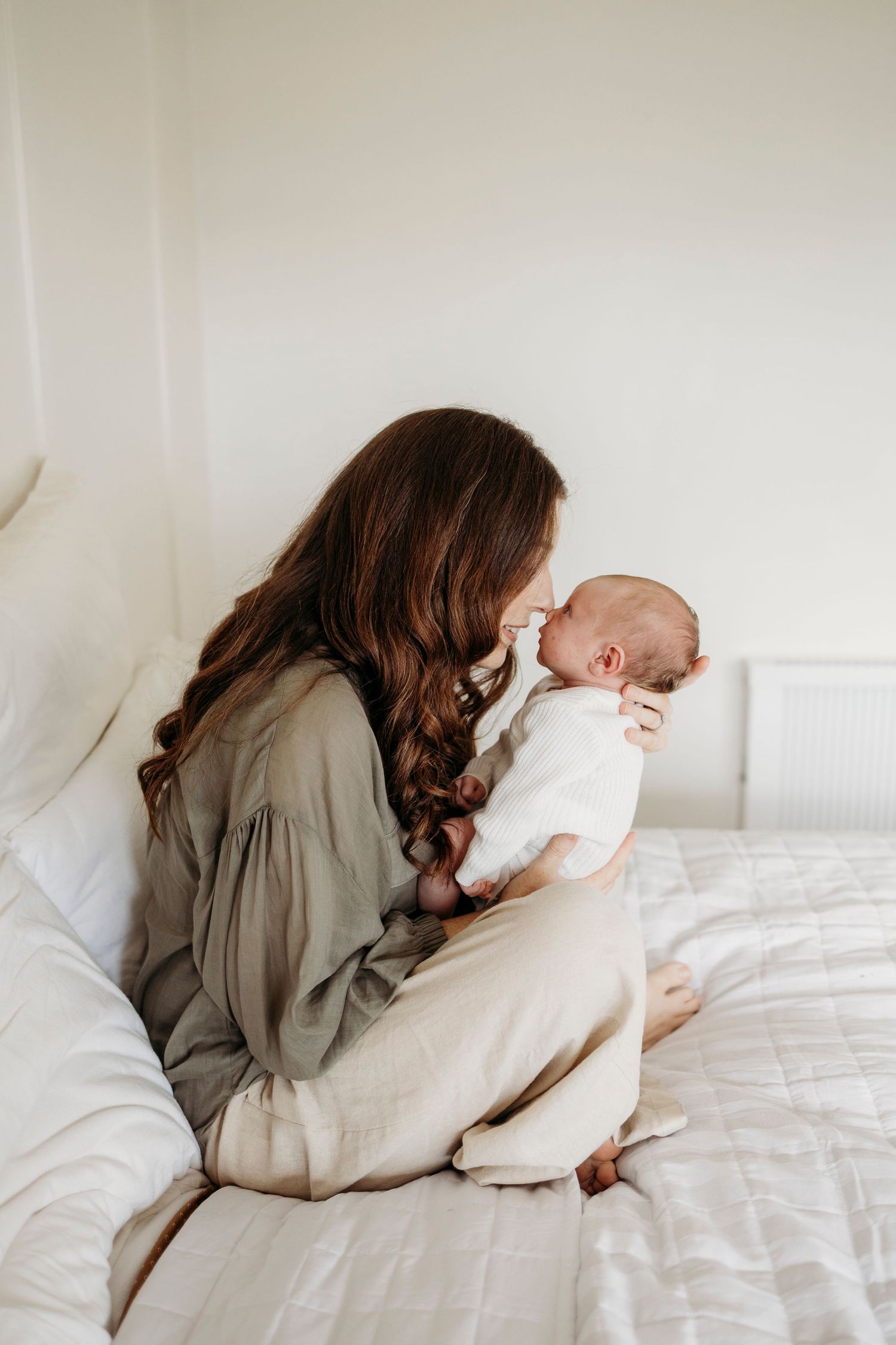 Brunette mother sitting on white bed kissing her newborn baby dressed in white.