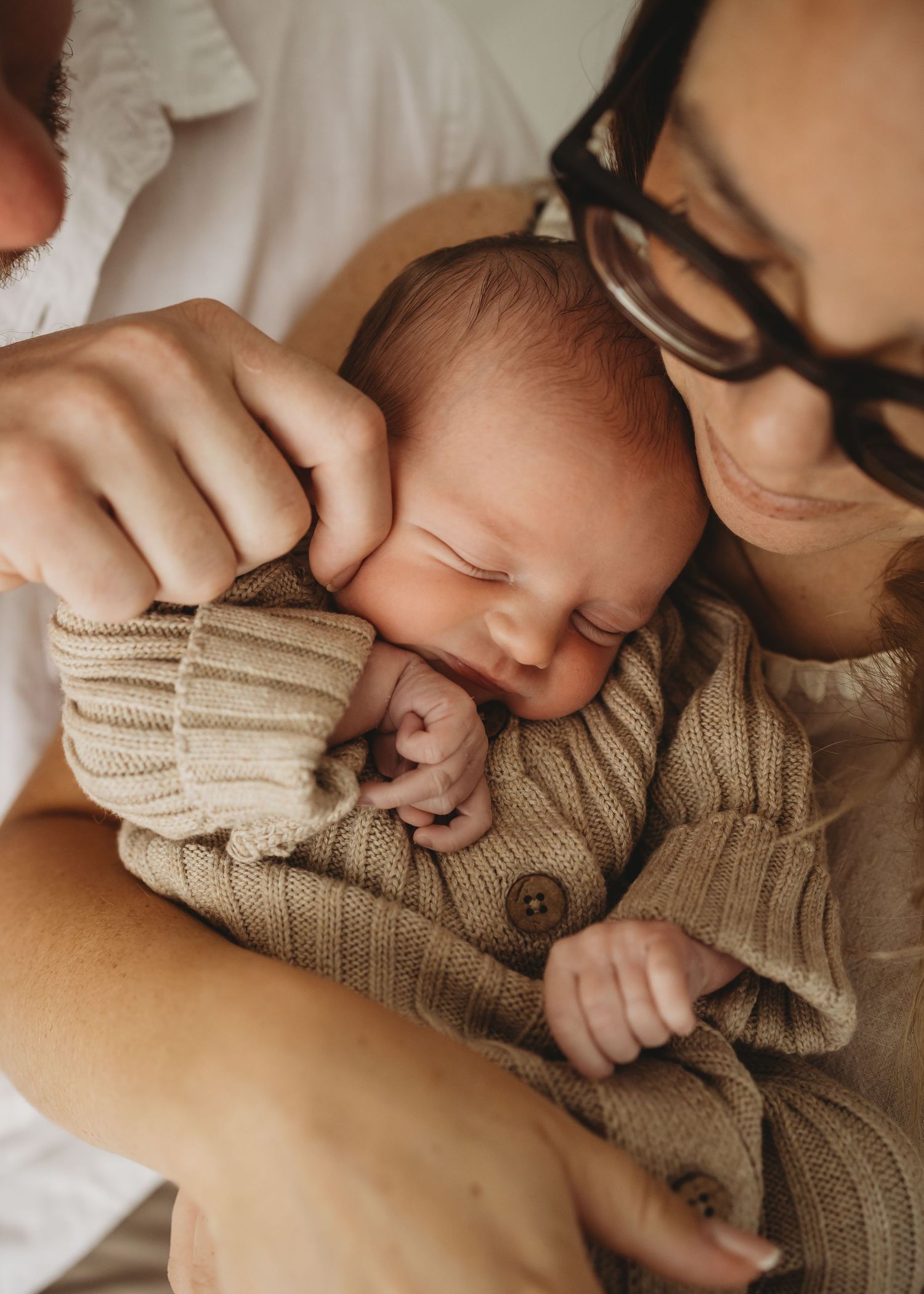 Close-up of sleeping newborn baby in knit outfit being gently held by parent with glasses.