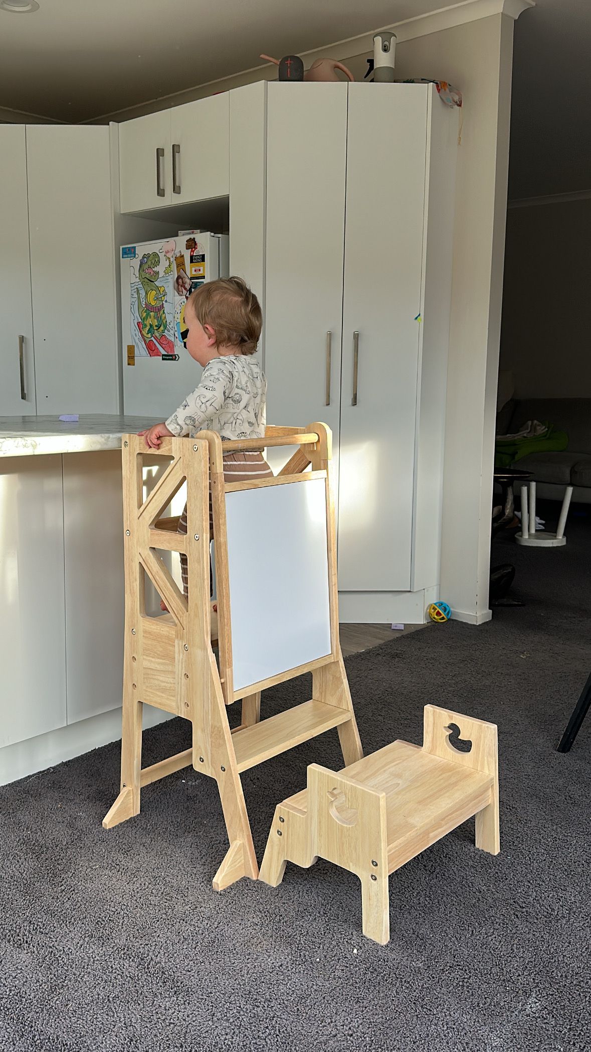 Toddler standing on wooden learning tower kitchen helper next to white kitchen bench and cabinets.