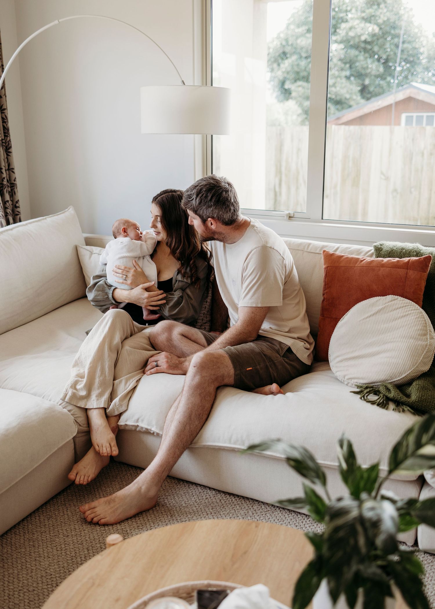 Loving couple sitting on white sofa with newborn baby in bright living room.