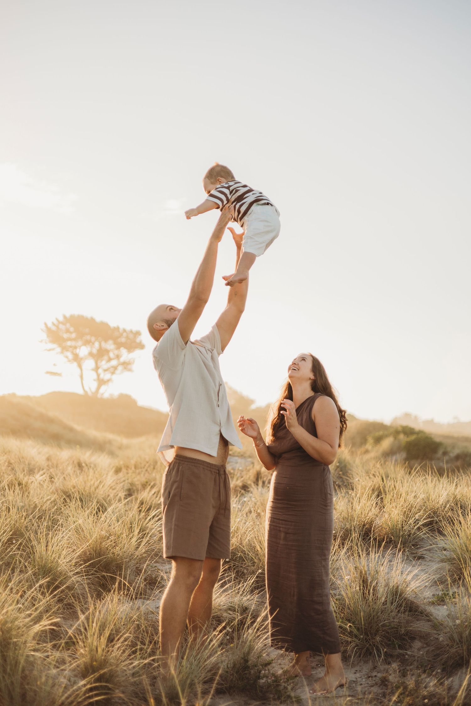 Father lifting baby high in air while mother watches in golden grassy beach dunes.
