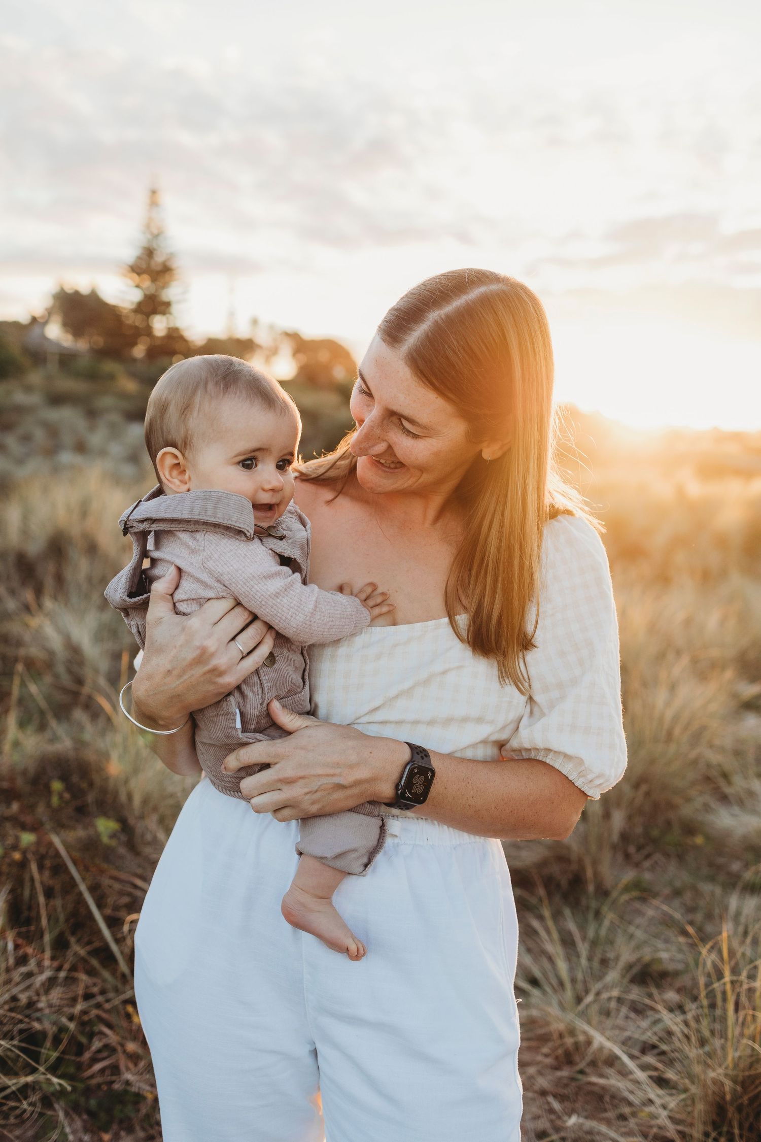 Mother holding smiling baby in golden sunset field wearing white outfit.