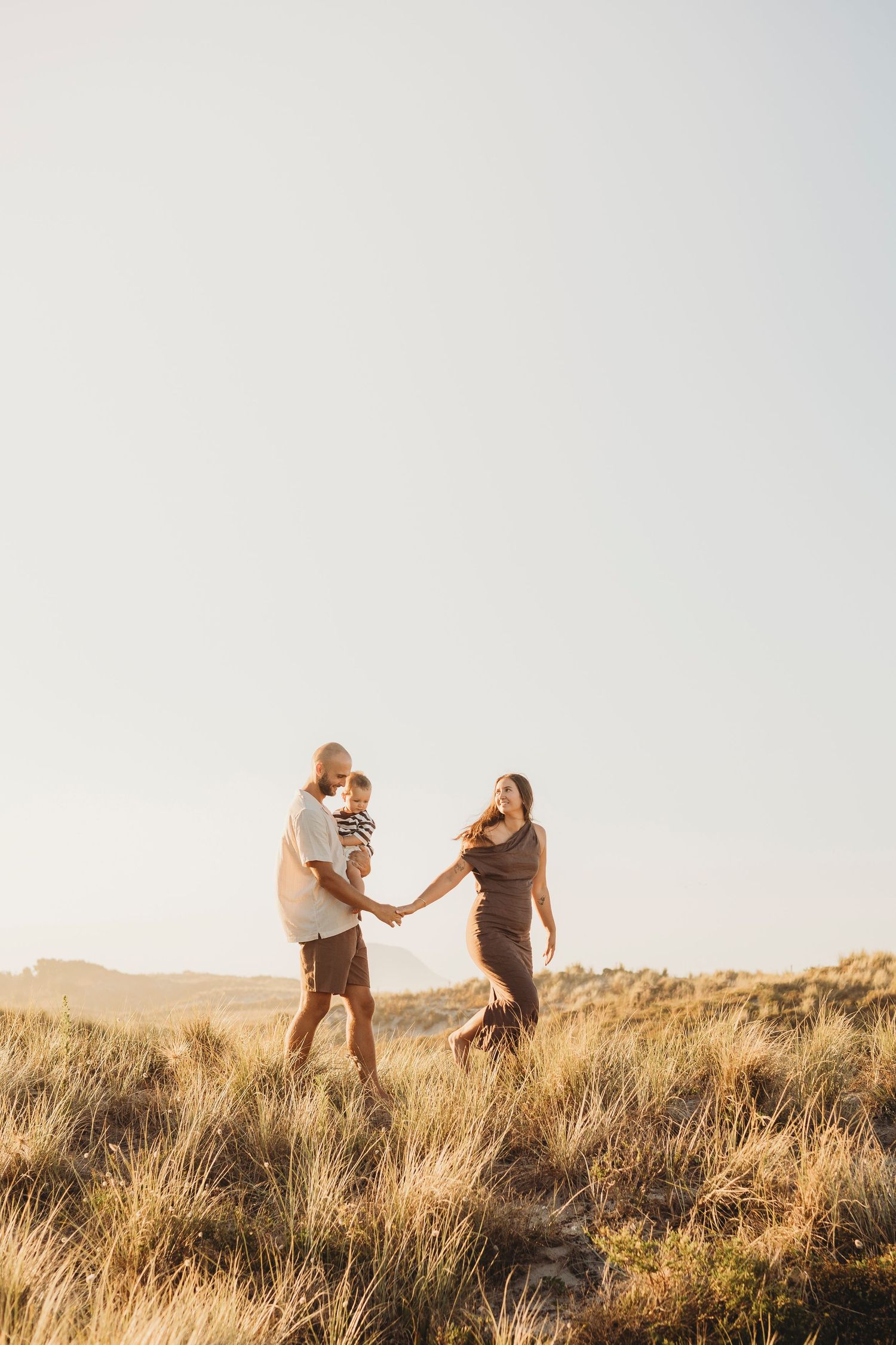 Family of three holding hands and dancing in golden grassy beach dunes at sunset.