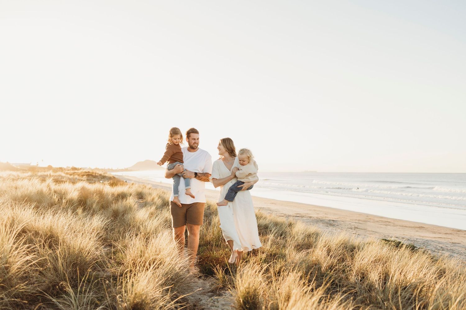 Parents holding two small children among golden beach dunes with ocean visible in background.