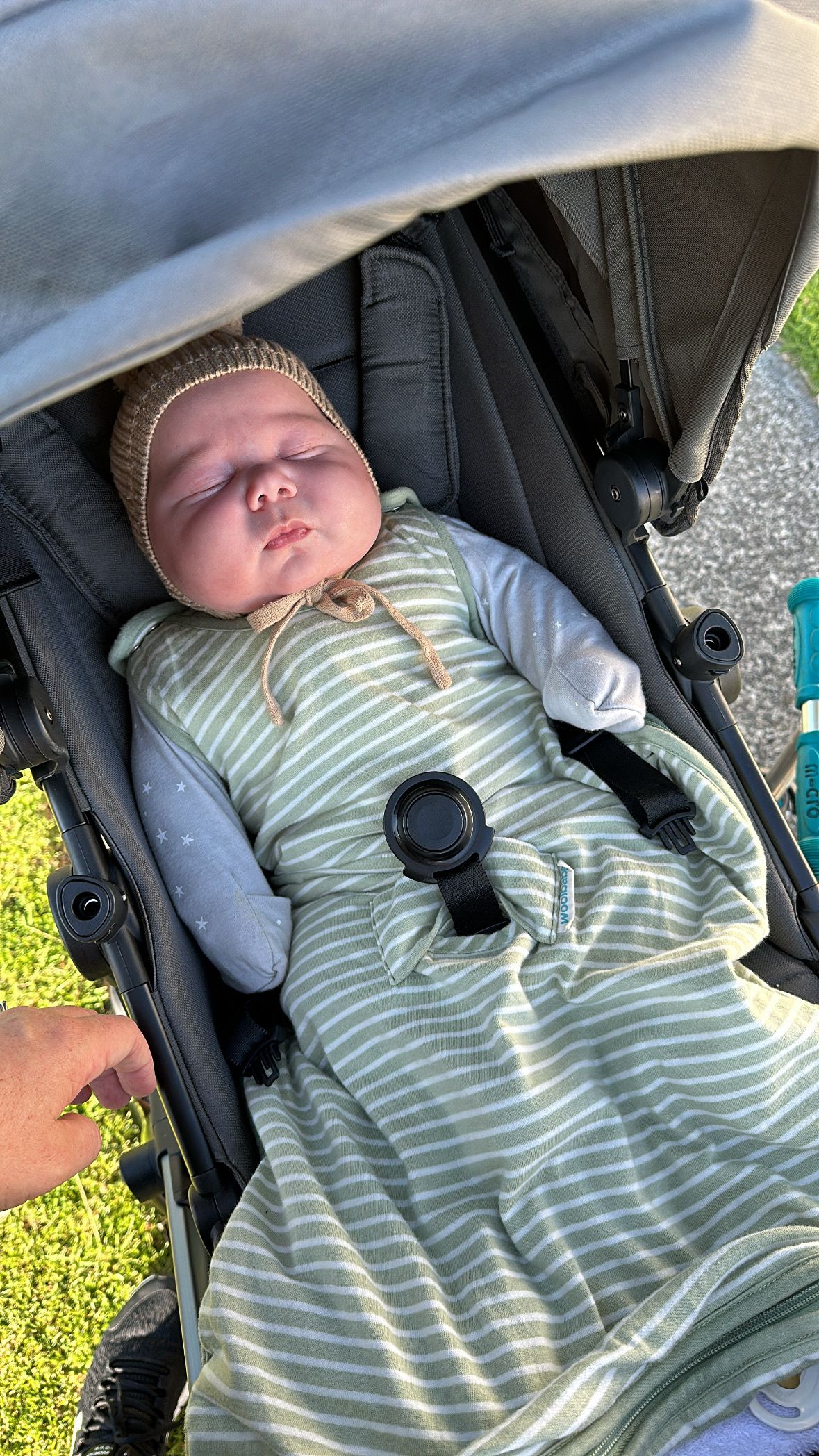Sleeping newborn baby in green striped sleeping bag secured in grey pram outdoors in sunlight.