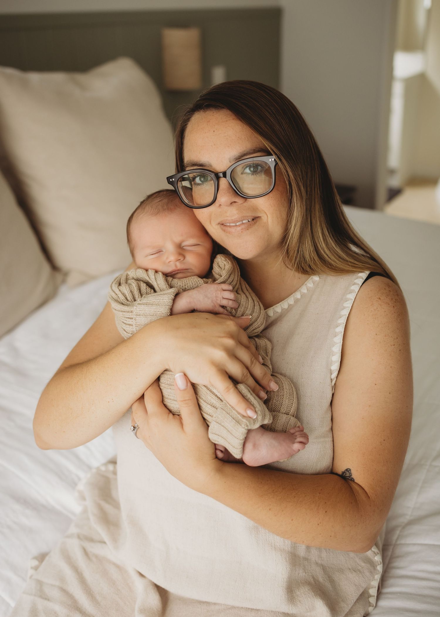 New mother wearing glasses tenderly holding sleeping newborn baby, warm indoor lifestyle portrait.