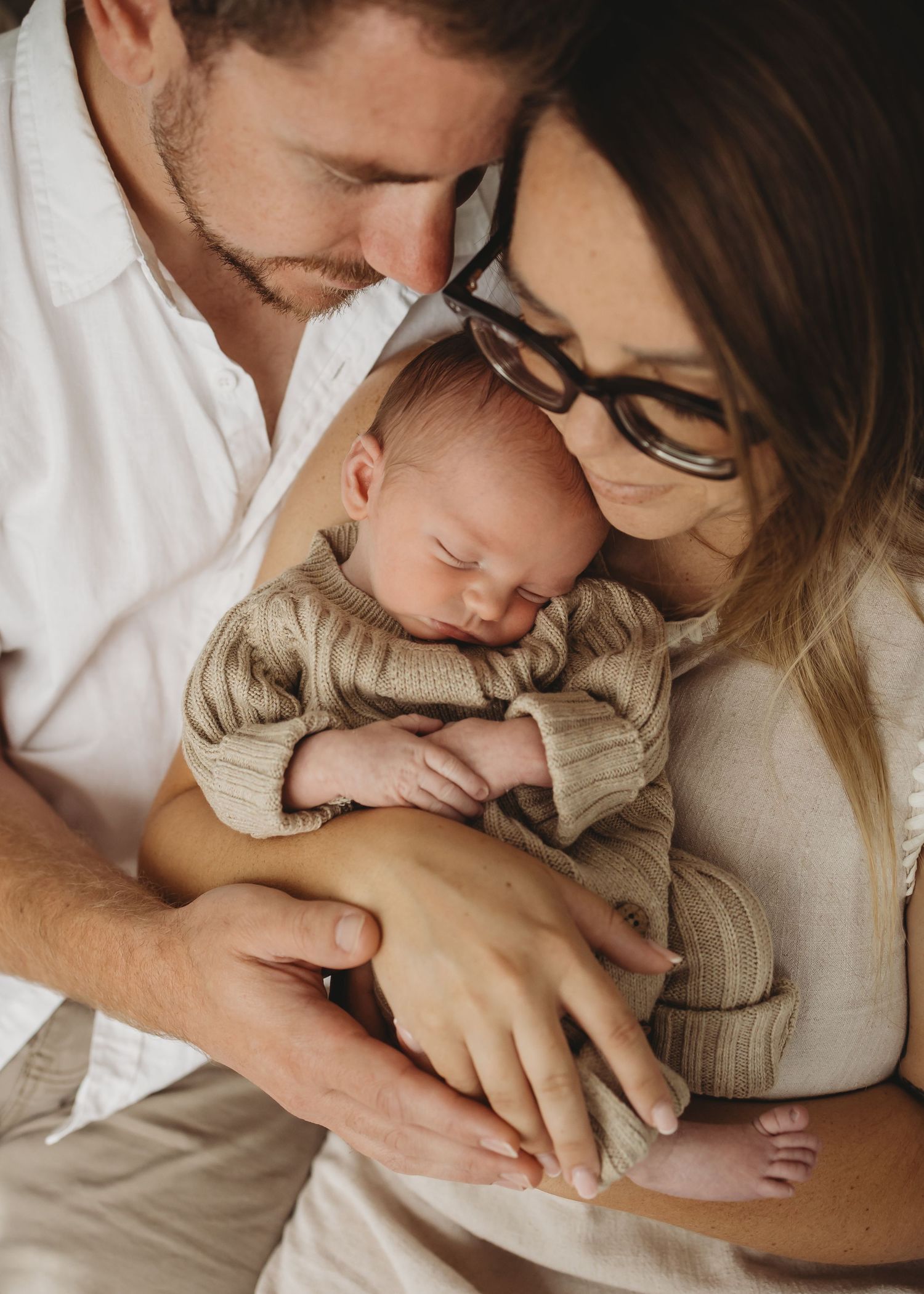 Loving parents holding their sleeping newborn baby dressed in a beige knit outfit.