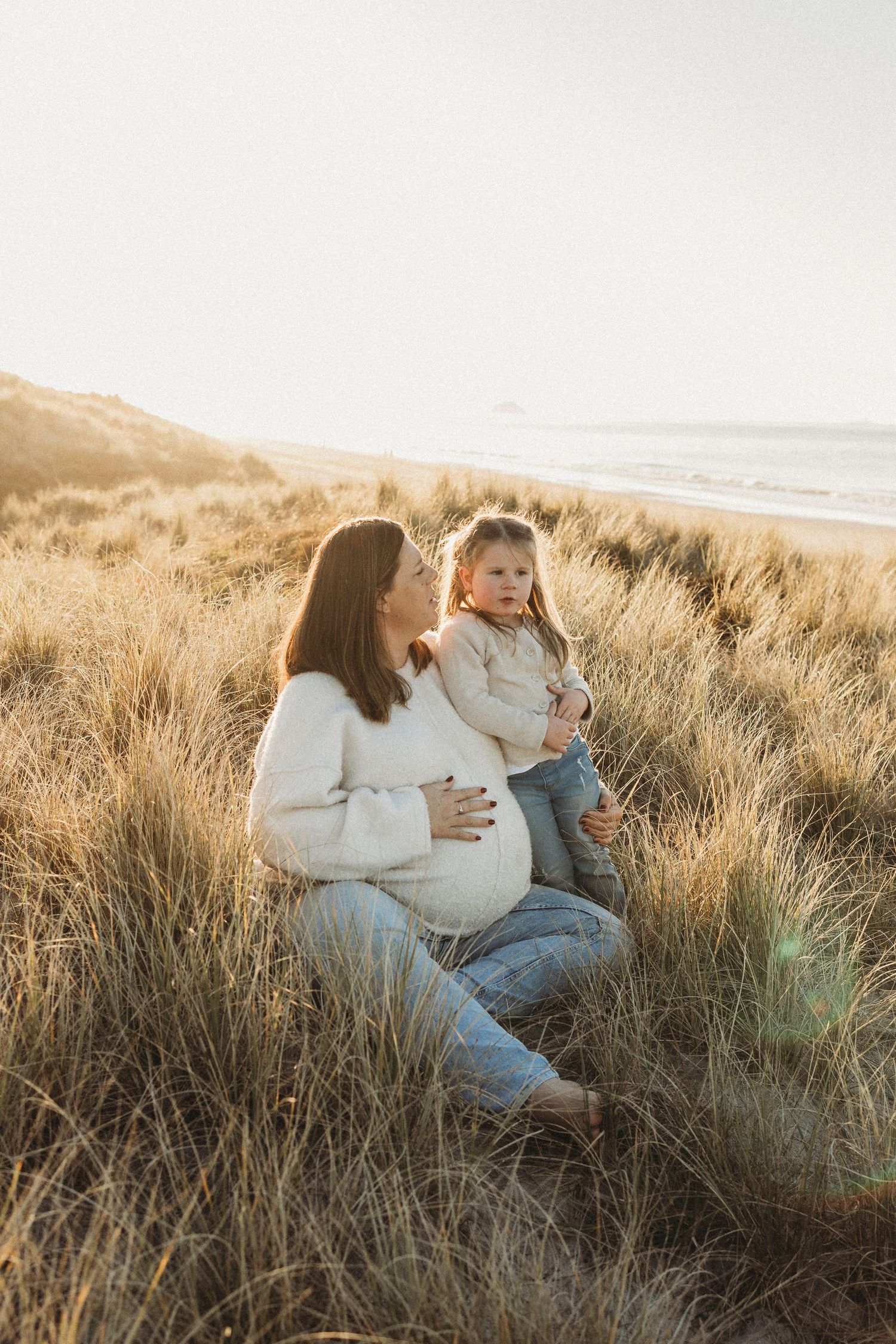 Pregnant woman sitting in golden beach dunes with toddler at sunset.