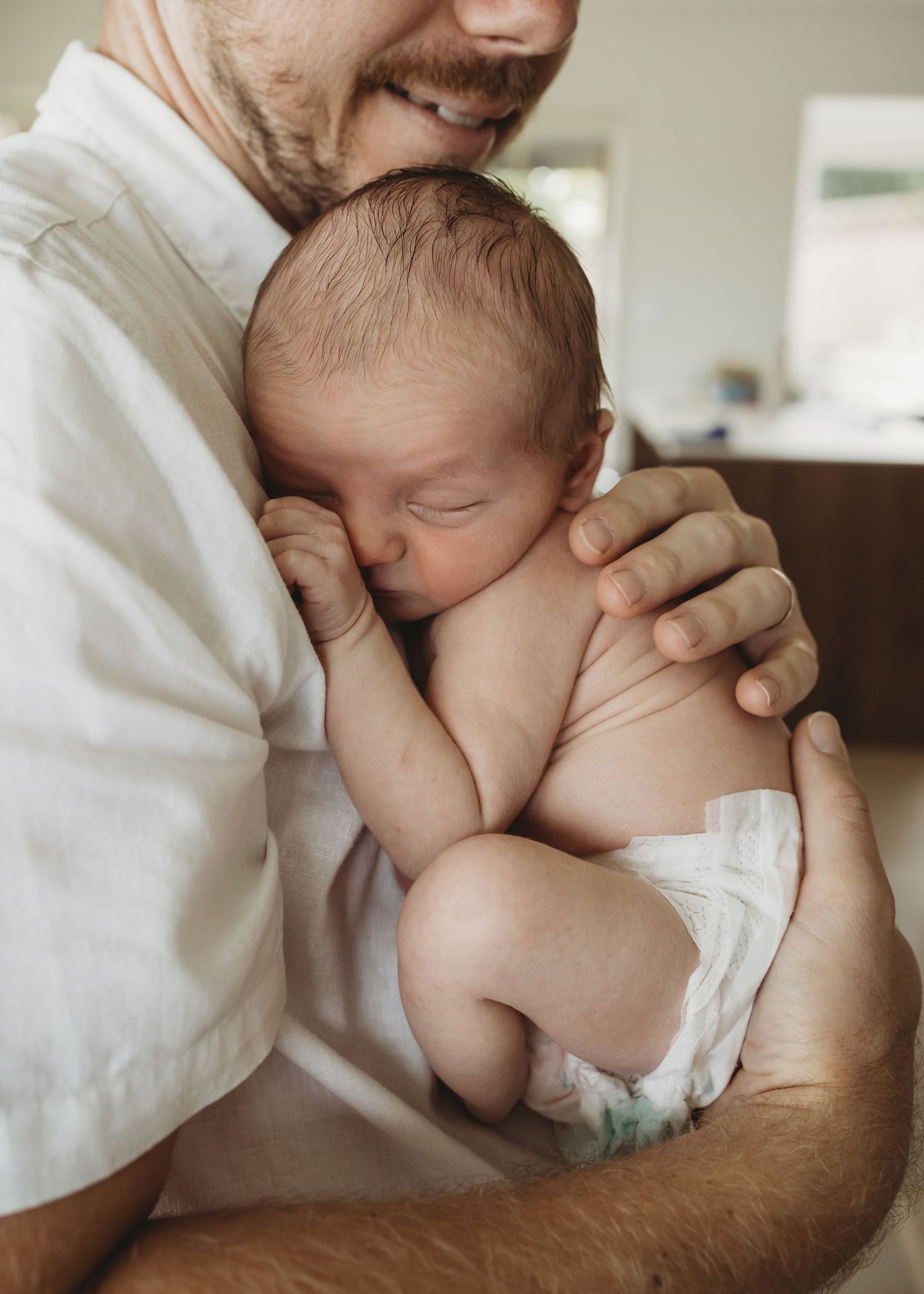 Father in white shirt tenderly holding sleeping newborn baby against his chest.